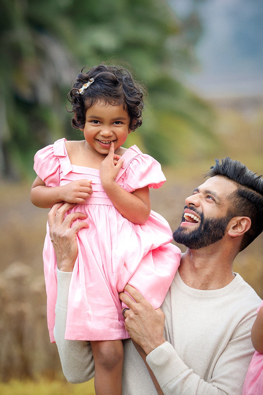 A man lifting a young girl in a pink dress outdoors, smiling at each other.