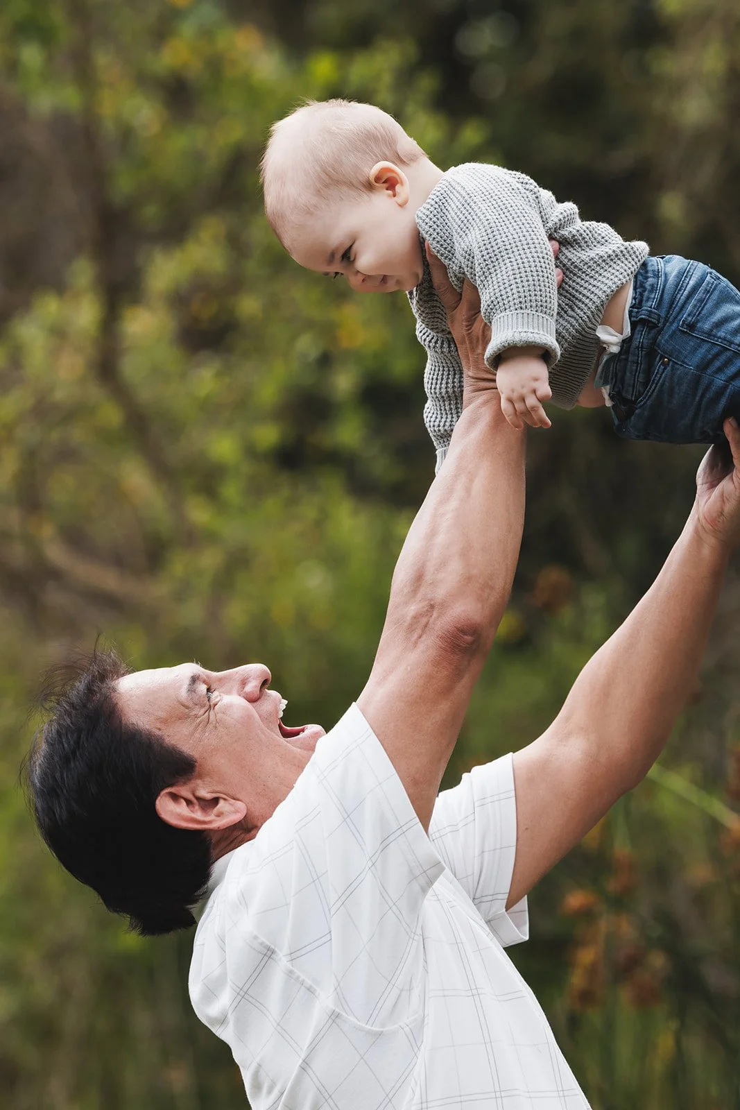 An elderly man lifting a young child in the air outdoors with trees and greenery in the background.