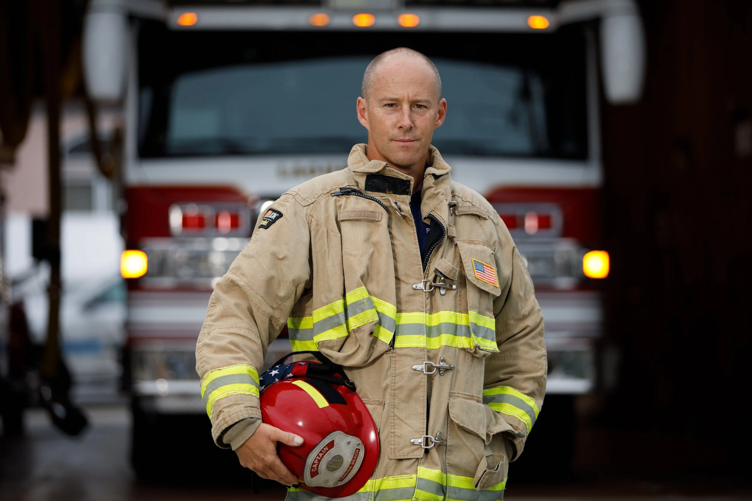 A firefighter in uniform holding a red helmet standing in front of a fire truck.