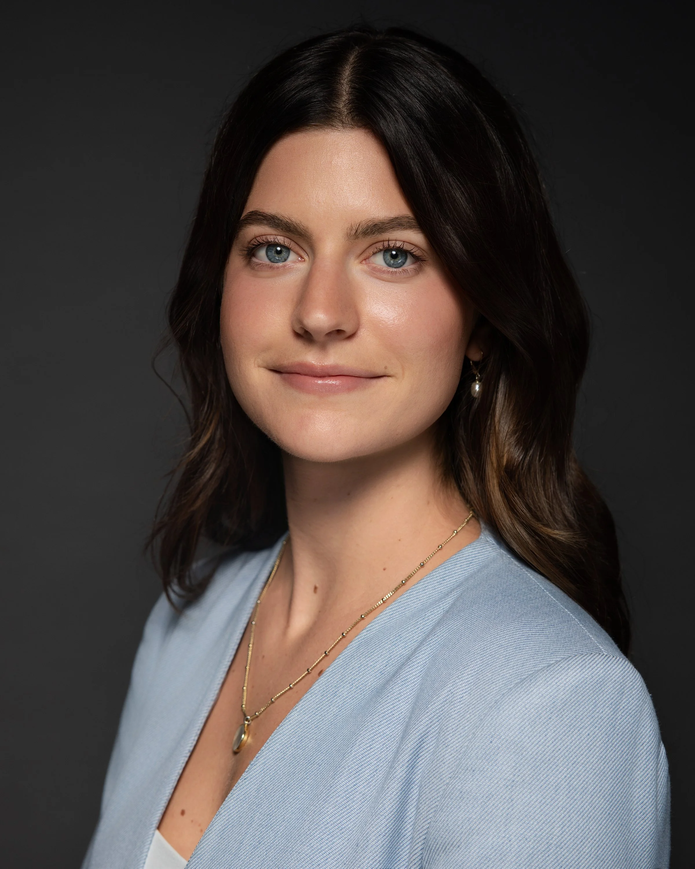 Portrait of a woman with dark brown hair, blue eyes, wearing a light blue blazer and gold jewelry, against a dark background.