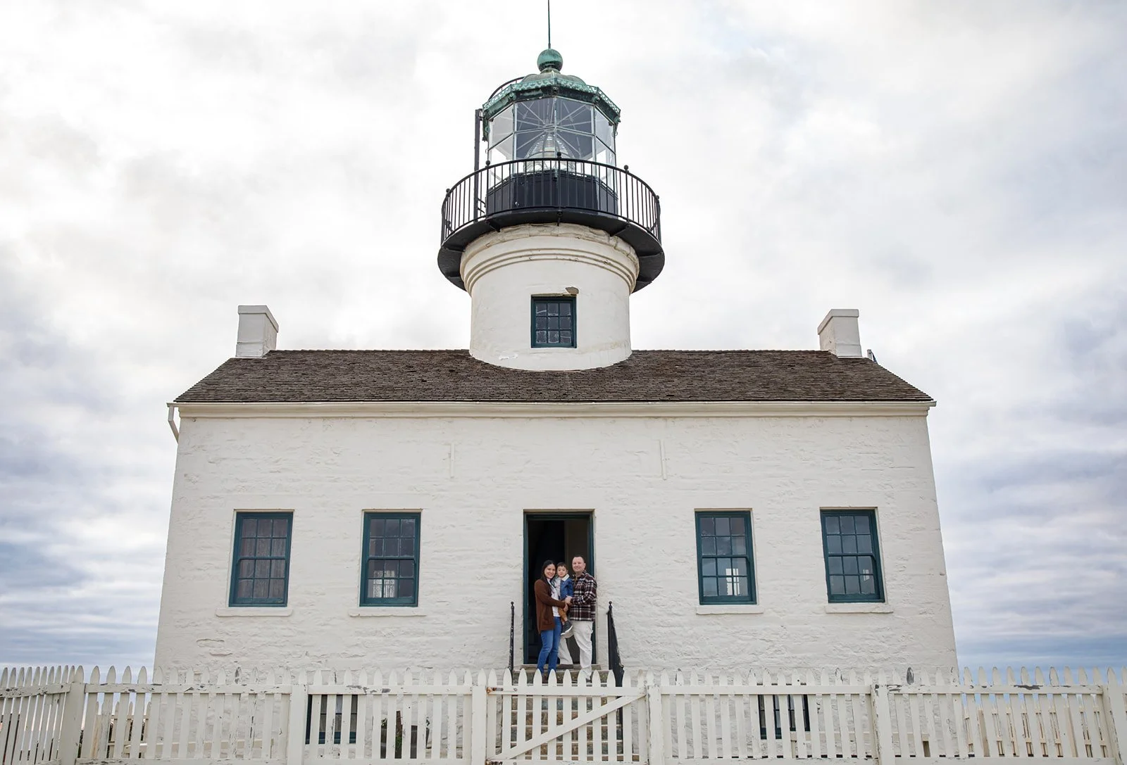 A white lighthouse with a balcony and glass enclosure on top, with a small window on the tower and four windows on the building's front. Four people are standing at the entrance.