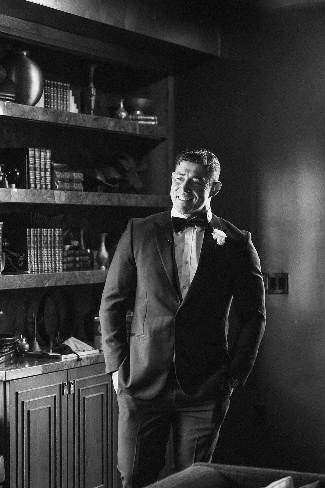 A man in a tuxedo with a boutonniere, smiling, standing in front of bookshelves.