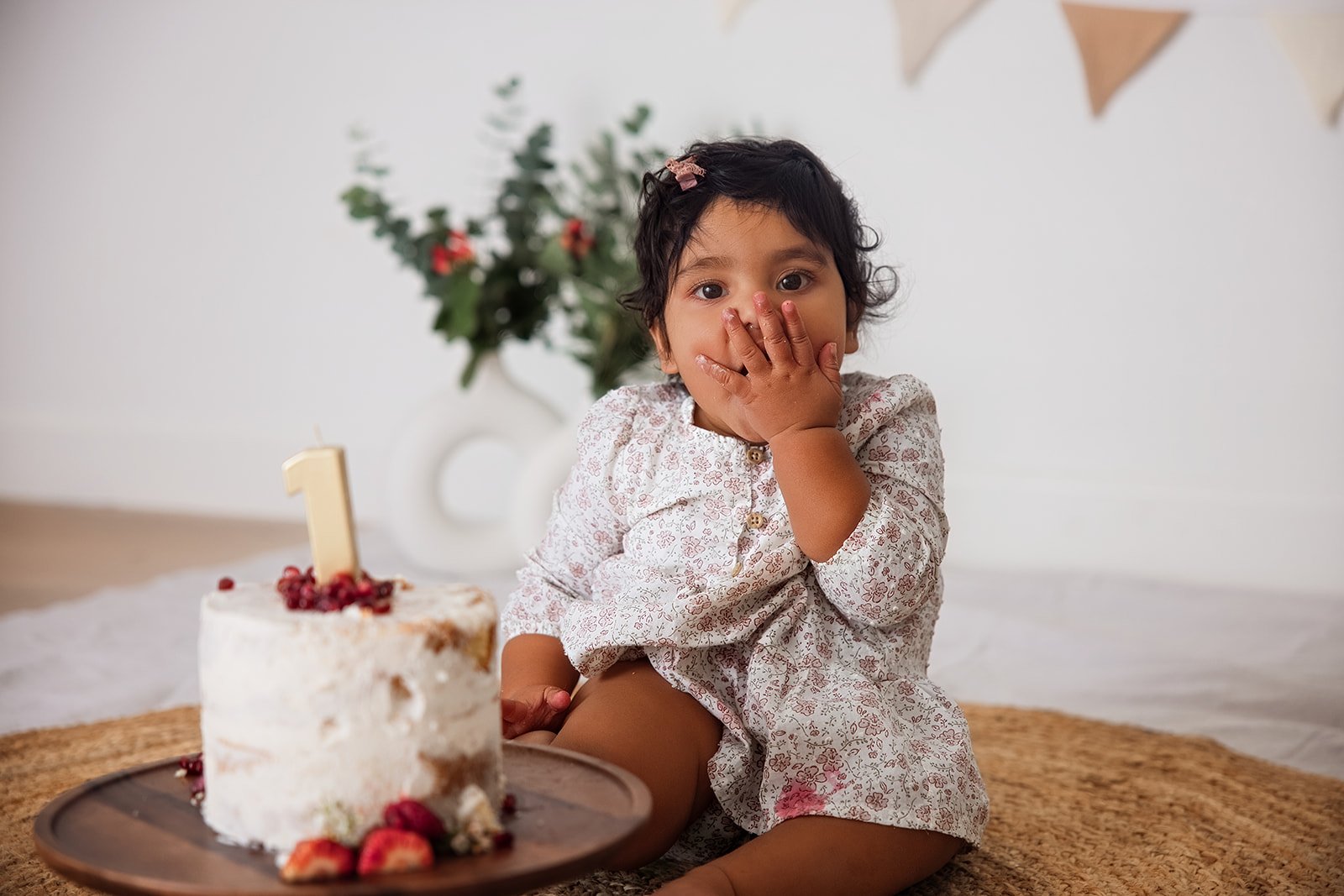 A young girl with dark curly hair and a pink hair clip covering her mouth with her hand, sitting on a woven mat in front of a birthday cake with a number one candle. There is a plant with red berries in the background and decorating banners overhead.