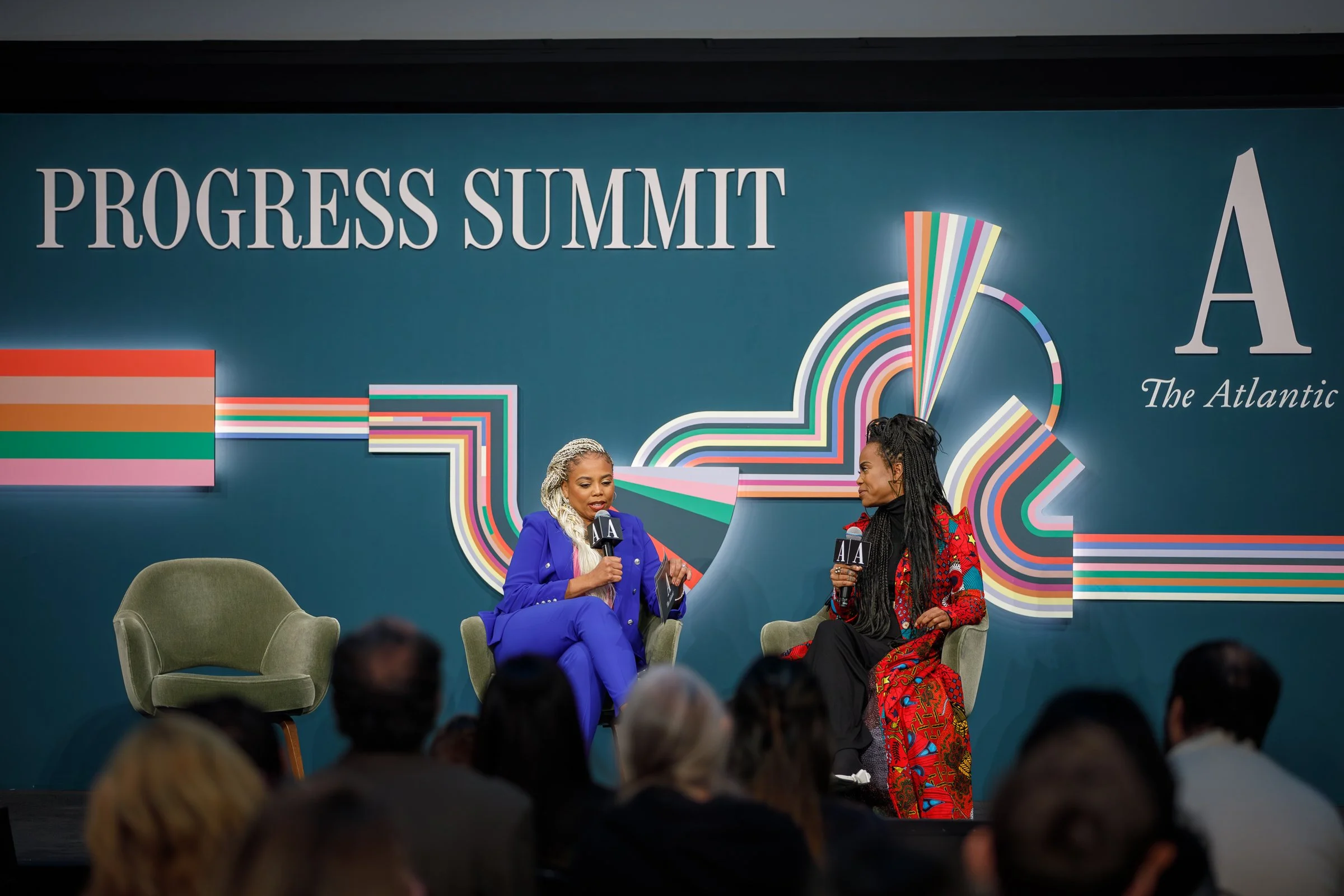 Two women speaking at the Progress Summit, sitting on chairs and holding microphones with 'A' logos, in front of a teal backdrop with colorful graphic designs and the text 'PROGRESS SUMMIT'.
