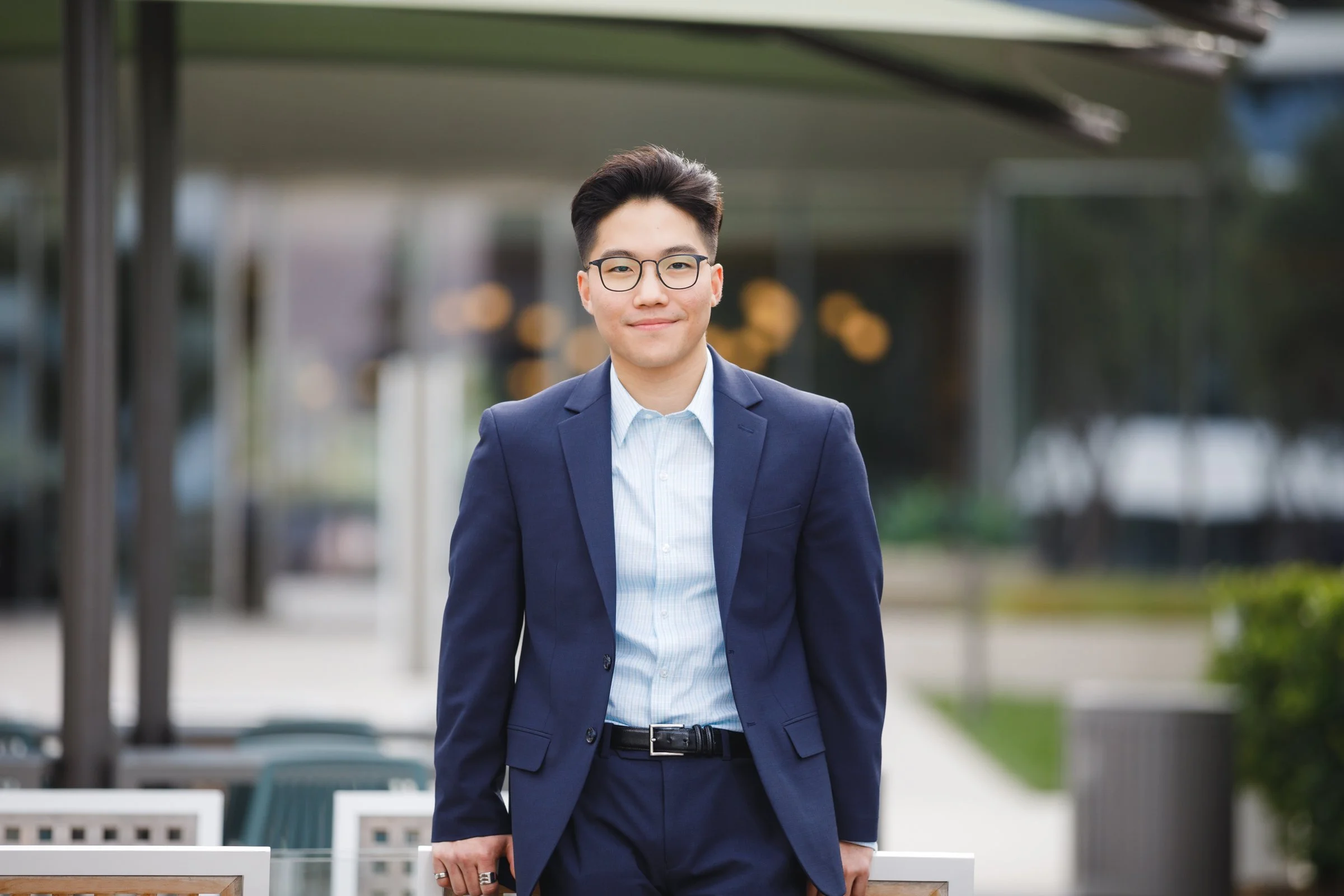 A young man wearing glasses and a navy blue suit, standing outdoors with a slight smile.