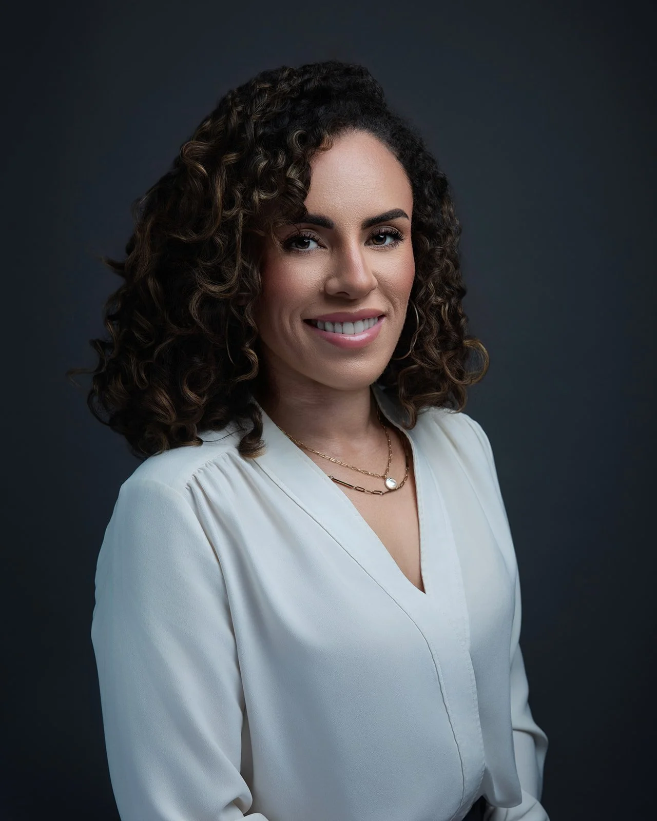 A woman with curly brown hair smiling in front of a dark background, wearing a white blouse and layered gold necklaces.