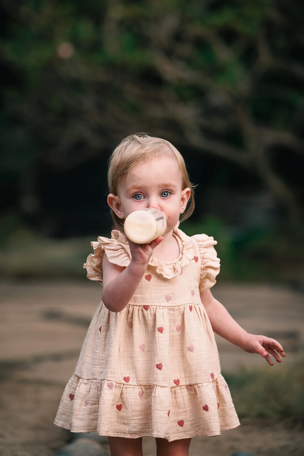 A young girl with blue eyes and blonde hair in a beige dress with red hearts drinks from a baby bottle outdoors.