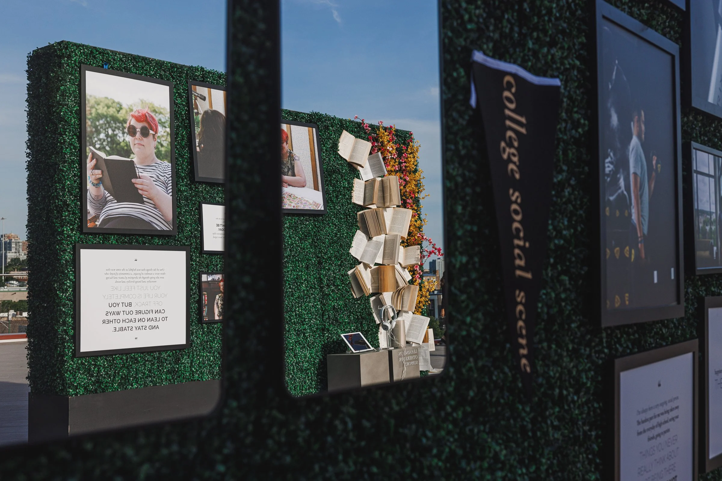 Outdoor photo of a display wall decorated with artificial greenery, framed photographs, and open books, with a city skyline and blue sky in the background.
