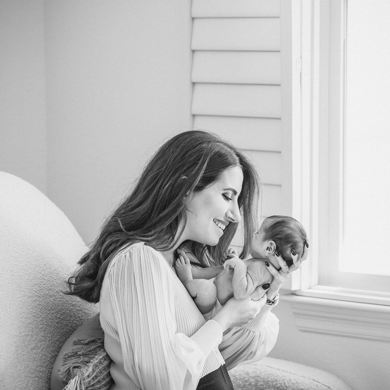 Black and white photo of a woman holding a newborn baby near a window, smiling and looking at the baby.