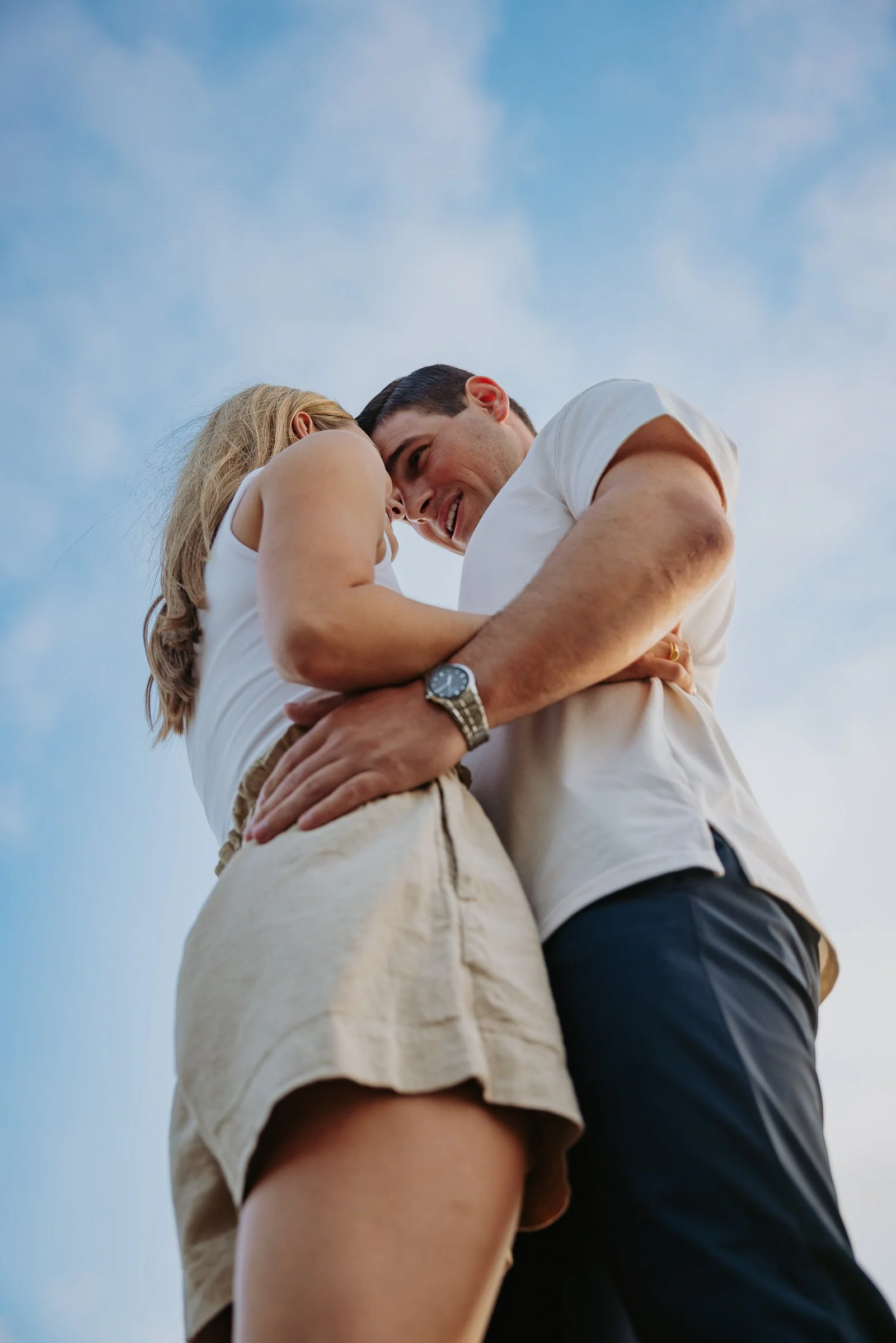 A smiling couple in casual clothing sharing an affectionate moment outdoors, viewed from below against a blue sky with some clouds.