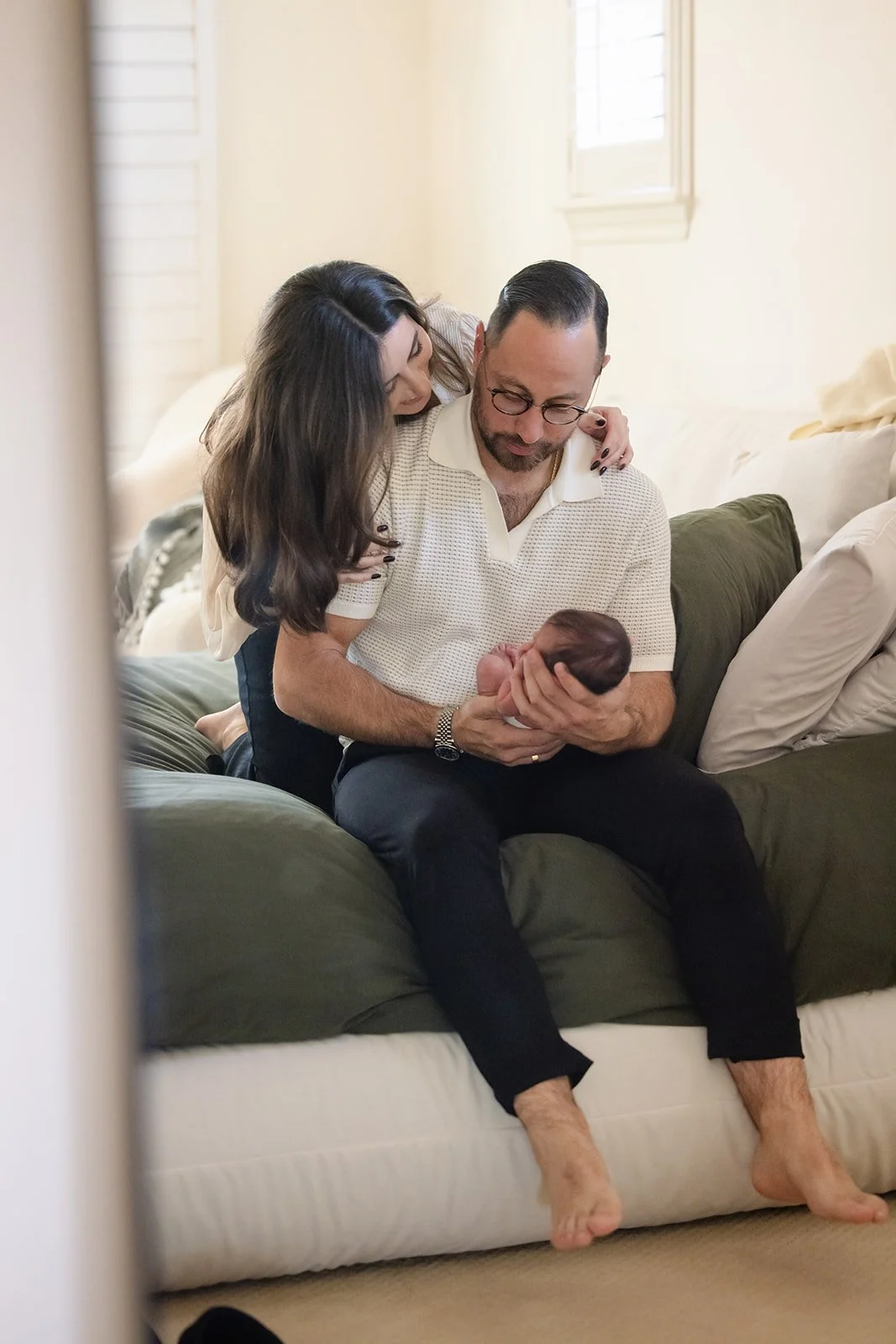A man sits on a couch holding a newborn baby, with a woman leaning over him and touching his shoulder. They appear to be in a cozy, well-lit living room.