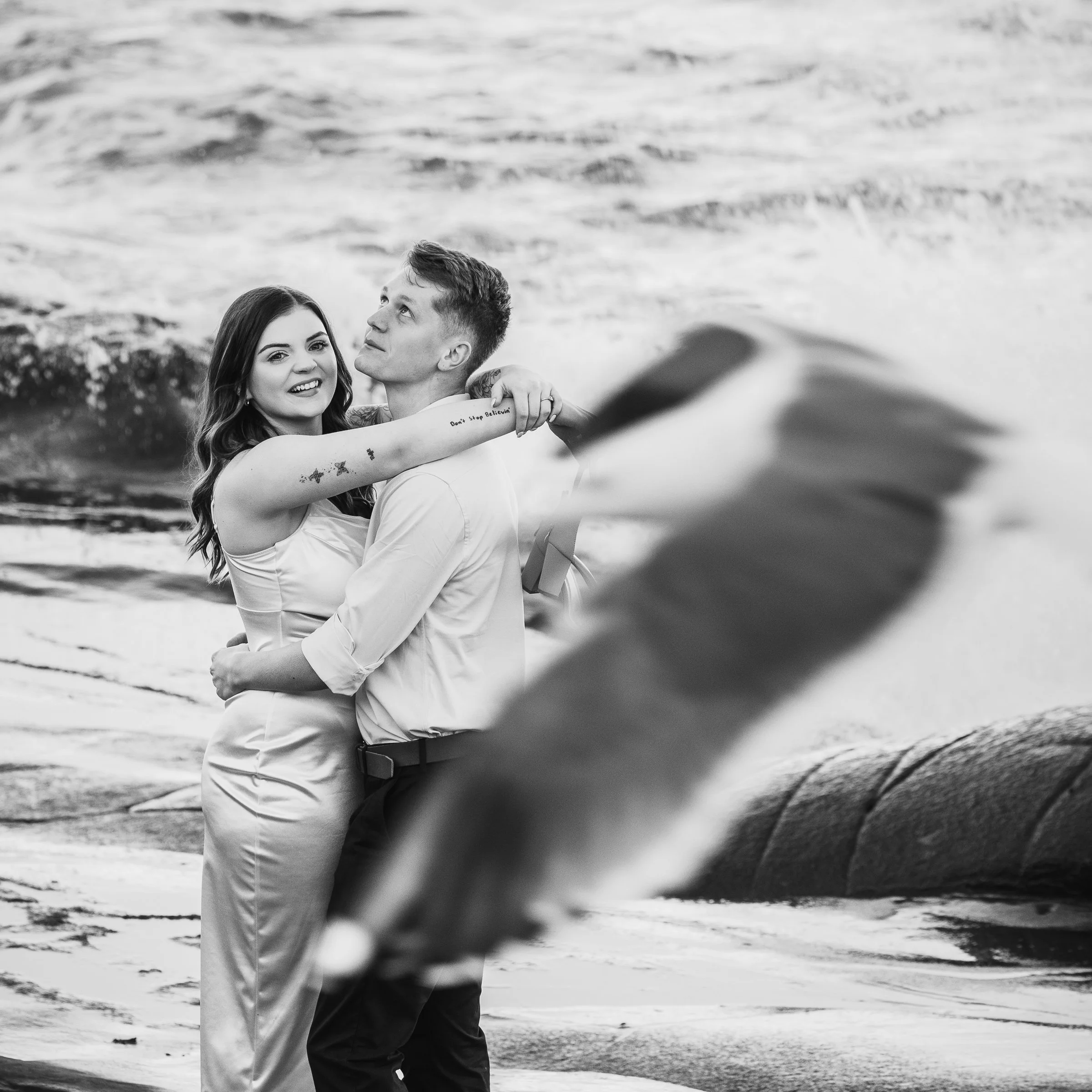 A black and white photo of a couple standing on a beach, embracing each other. The woman has long wavy hair and is smiling while looking at the camera, and the man is looking upwards. There is a blurred whale in the foreground and ocean waves in the 