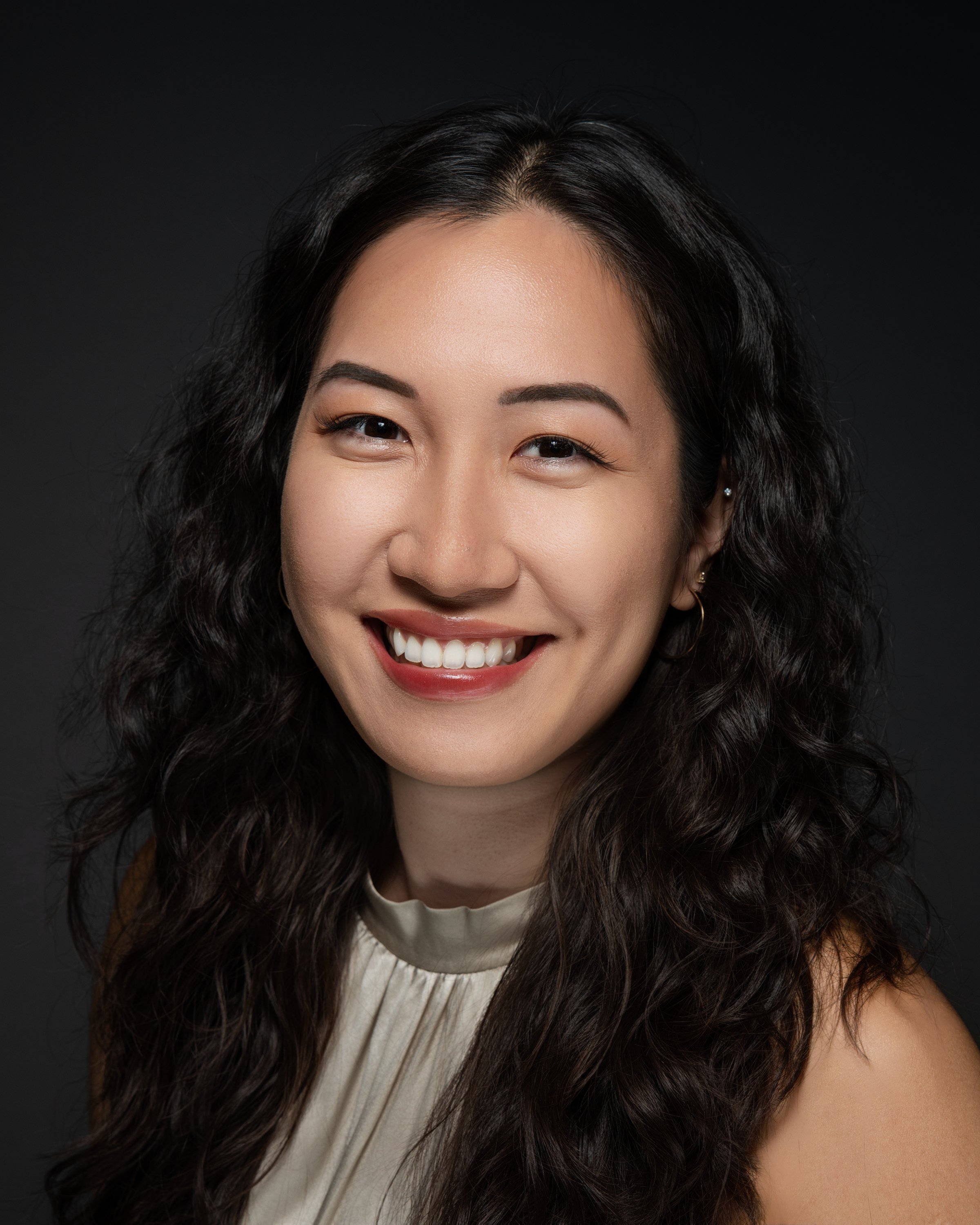 A smiling woman with long, wavy black hair, wearing a beige top, against a dark background.