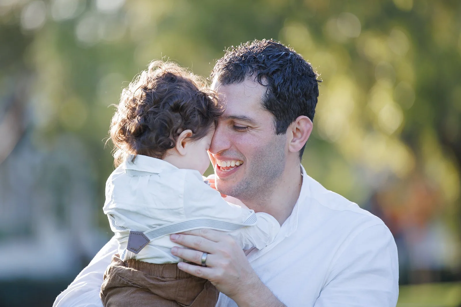 A man smiling and holding a young child close to his face outdoors.