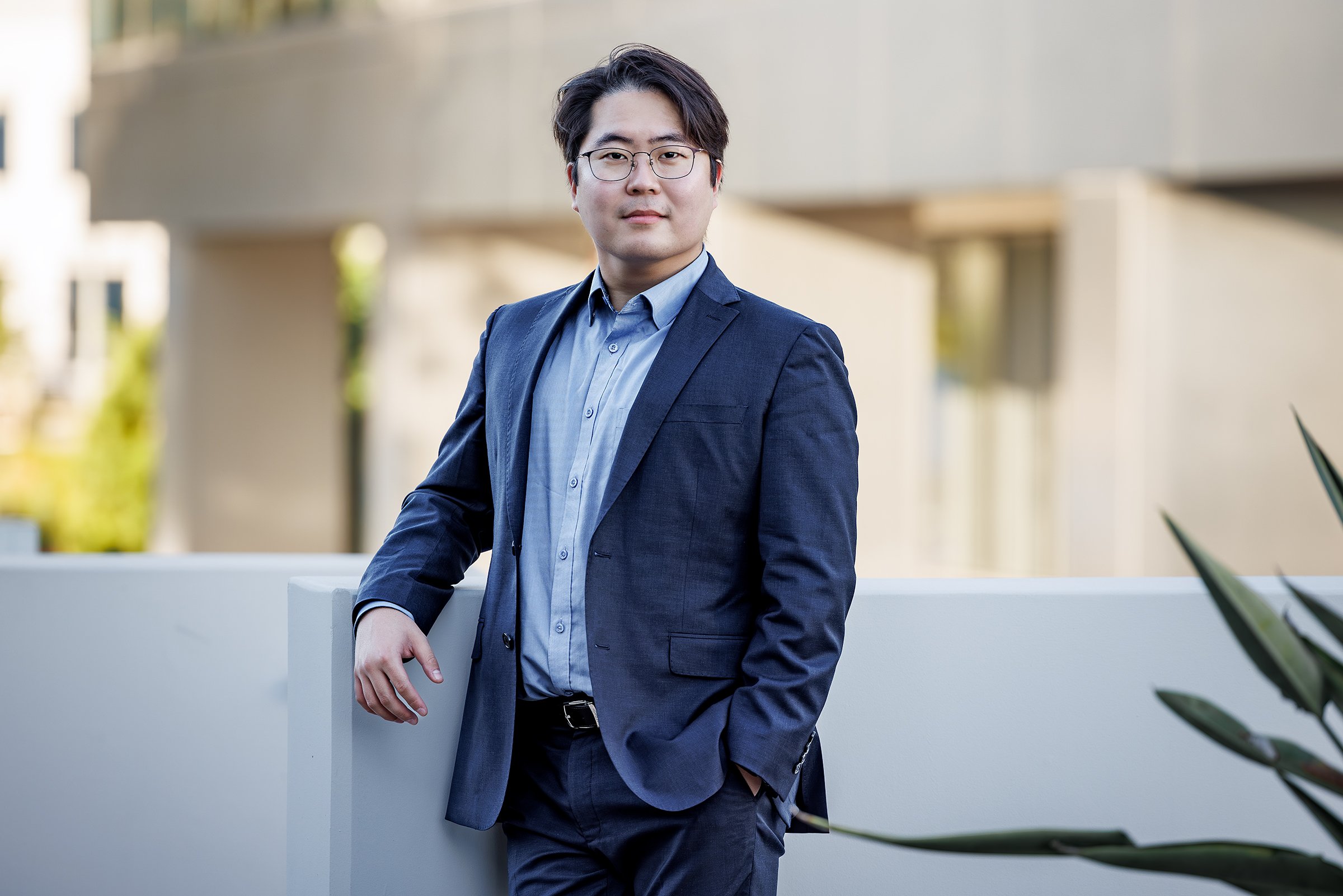 A man in a navy blue suit and glasses leaning on a white wall outdoors.