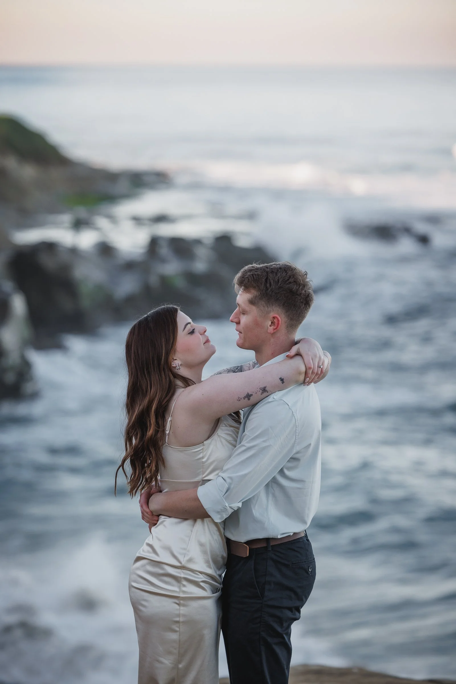 A couple hugging by the ocean during sunset, with the woman in a cream dress and long brown hair and the man in a white shirt and dark pants.