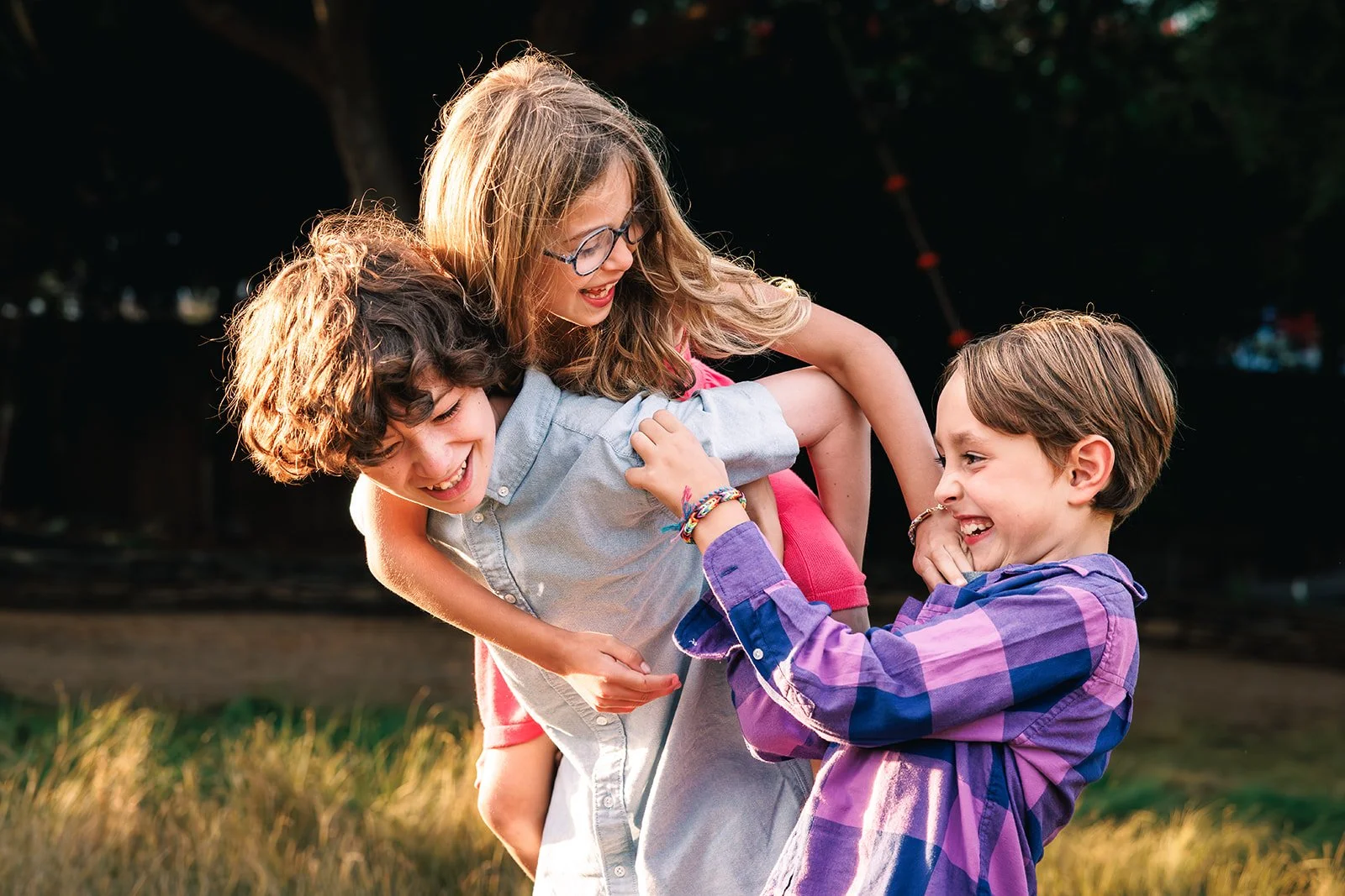 Three children, two boys and a girl, playing and laughing outdoors in a grassy area during sunset, engaging in a playful wrestling or tickling game.