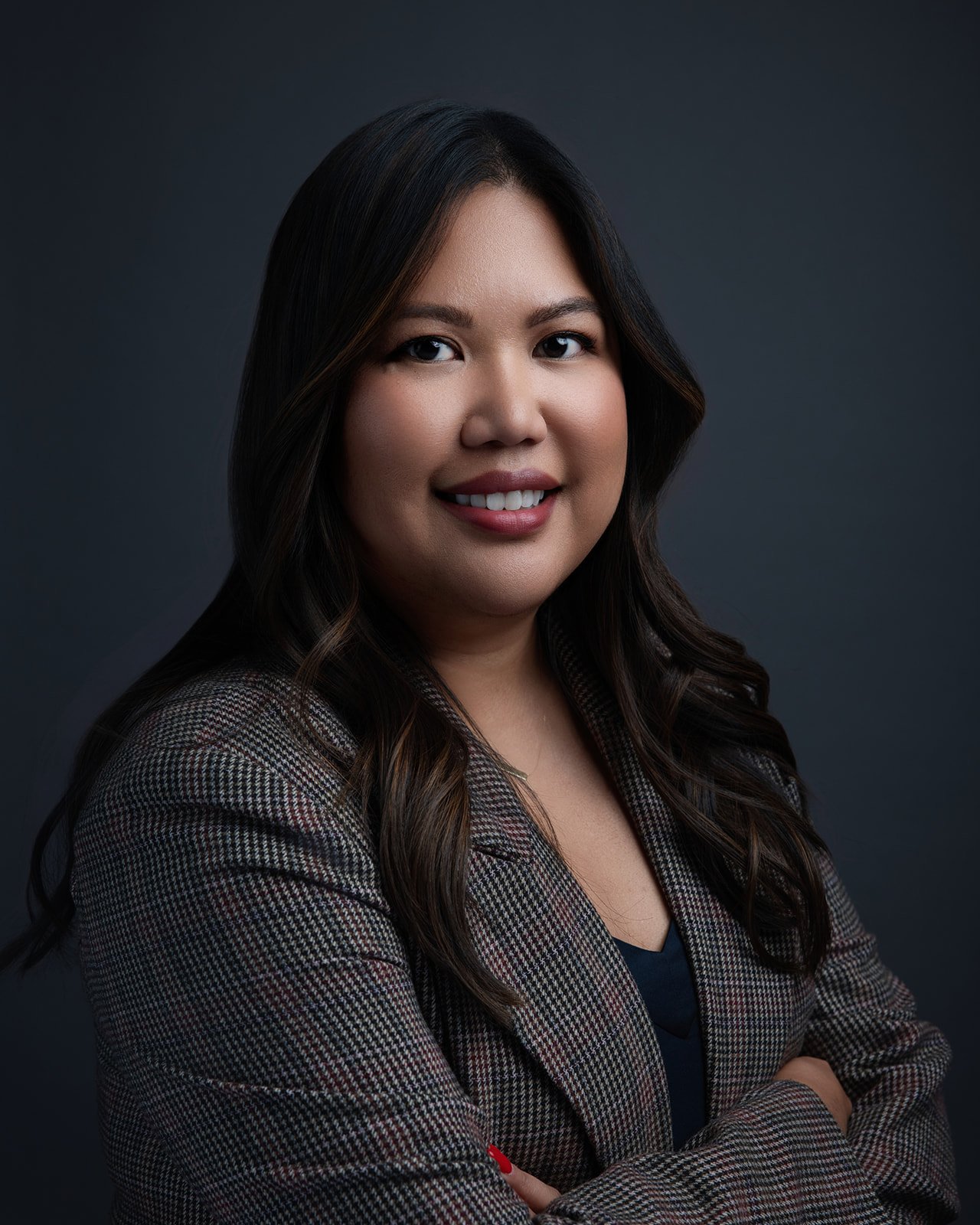 A professional woman with dark brown hair styled in loose waves, smiling with arms crossed, wearing a checkered blazer over a dark top, against a dark background.
