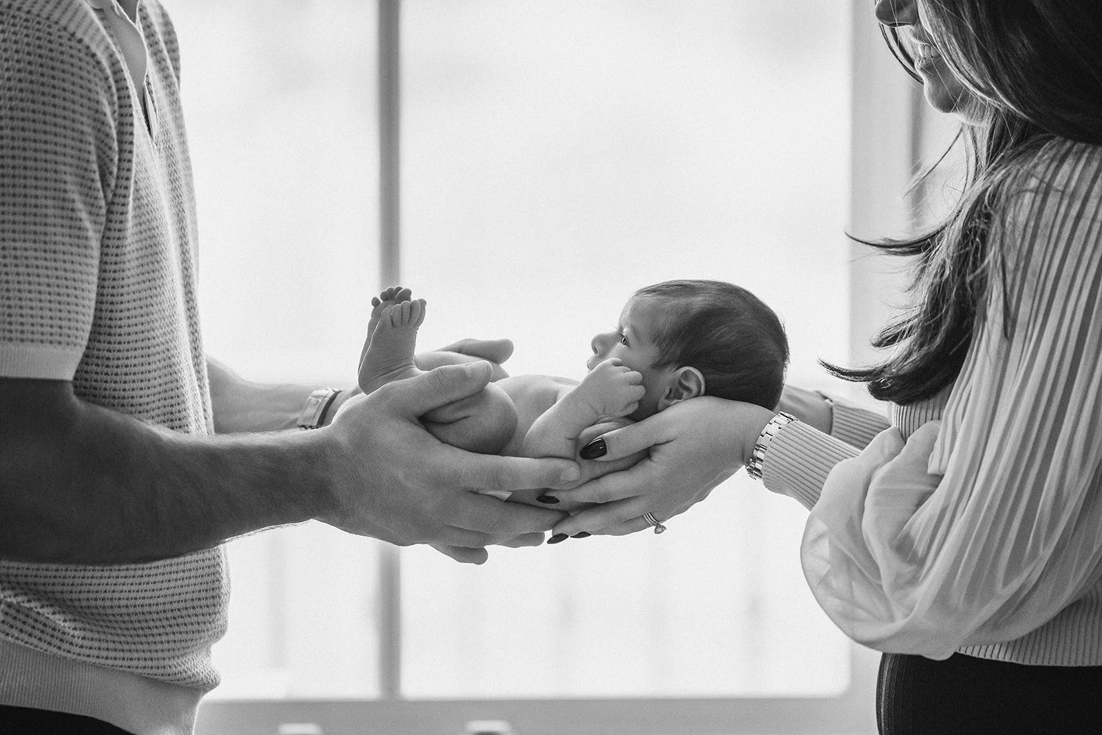 A newborn baby being held by two adults, one on each side, in a hospital or clinic setting.