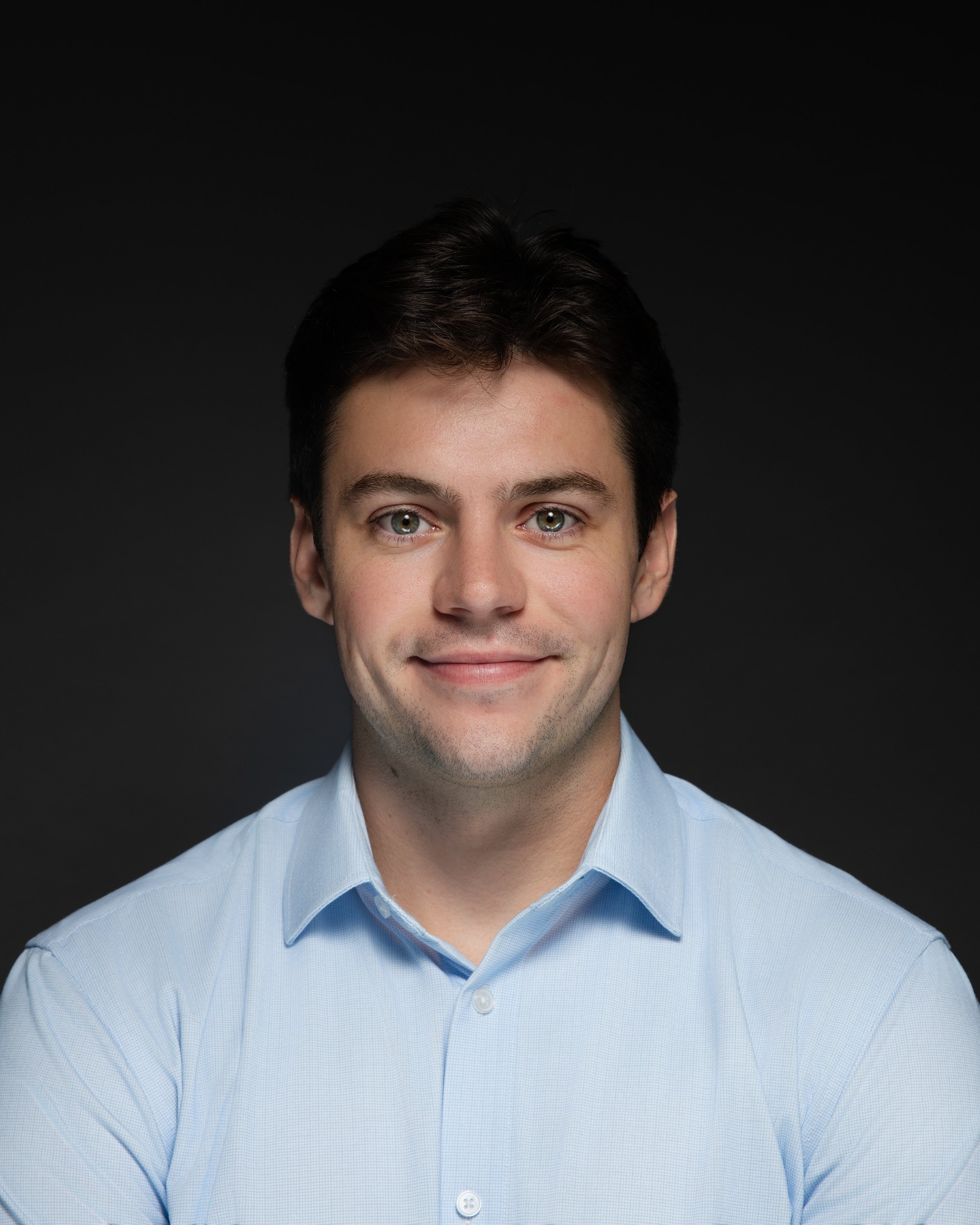 A young man with dark hair and light skin, wearing a light blue dress shirt, smiling against a dark background.