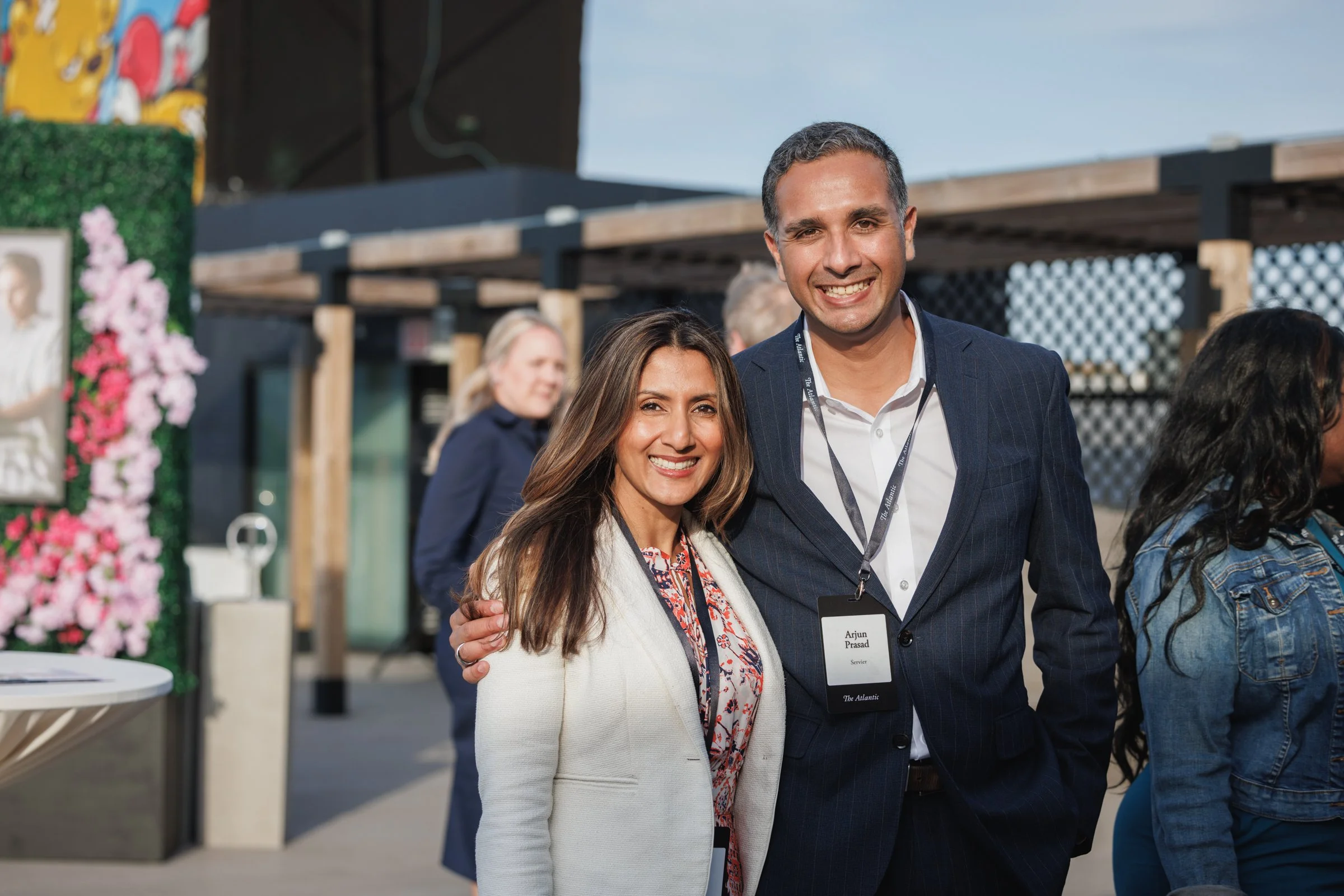 A man and woman smiling and standing close together outdoors at a professional event. The man is wearing a navy suit with a white shirt and a conference badge, and the woman is in a cream blazer with a patterned blouse. They are surrounded by other p
