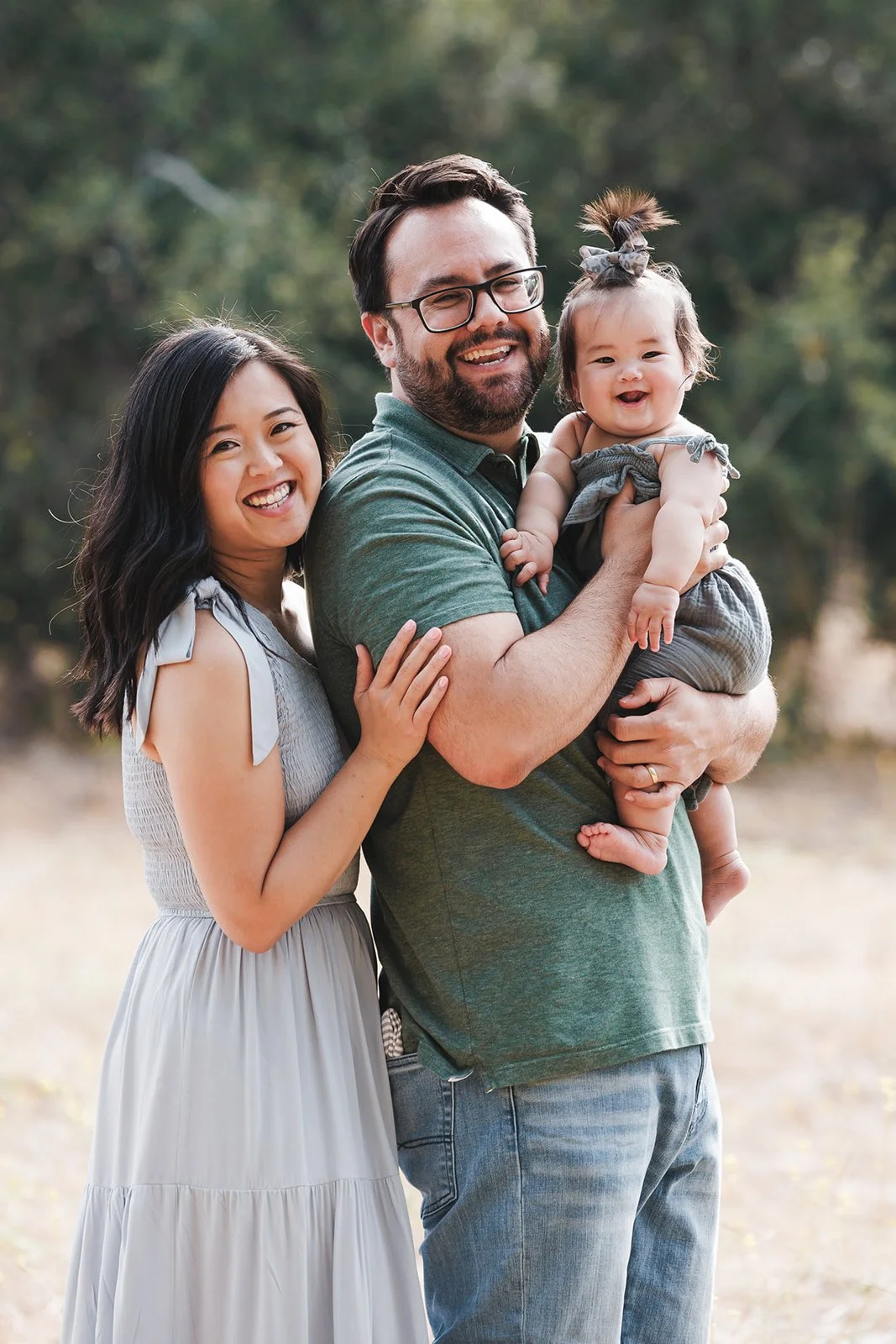 A happy family of three outdoors, a woman with long black hair, a man with glasses, beard, and short dark hair, holding a smiling baby girl with a gray bow in her hair, in a natural setting with trees in the background.