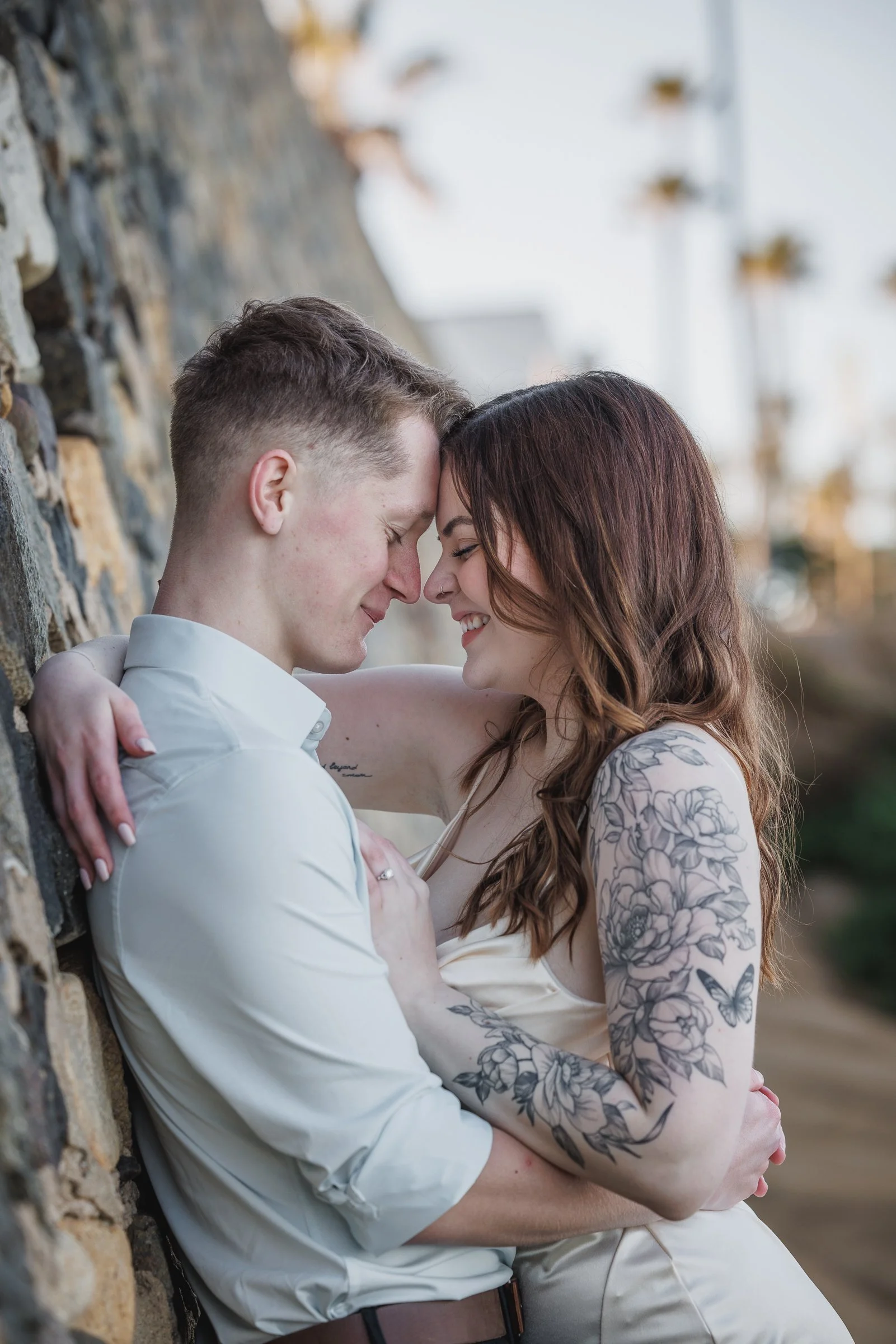 A young couple is leaning nose-to-nose against a stone wall, smiling with eyes closed, showing intimacy and affection.
