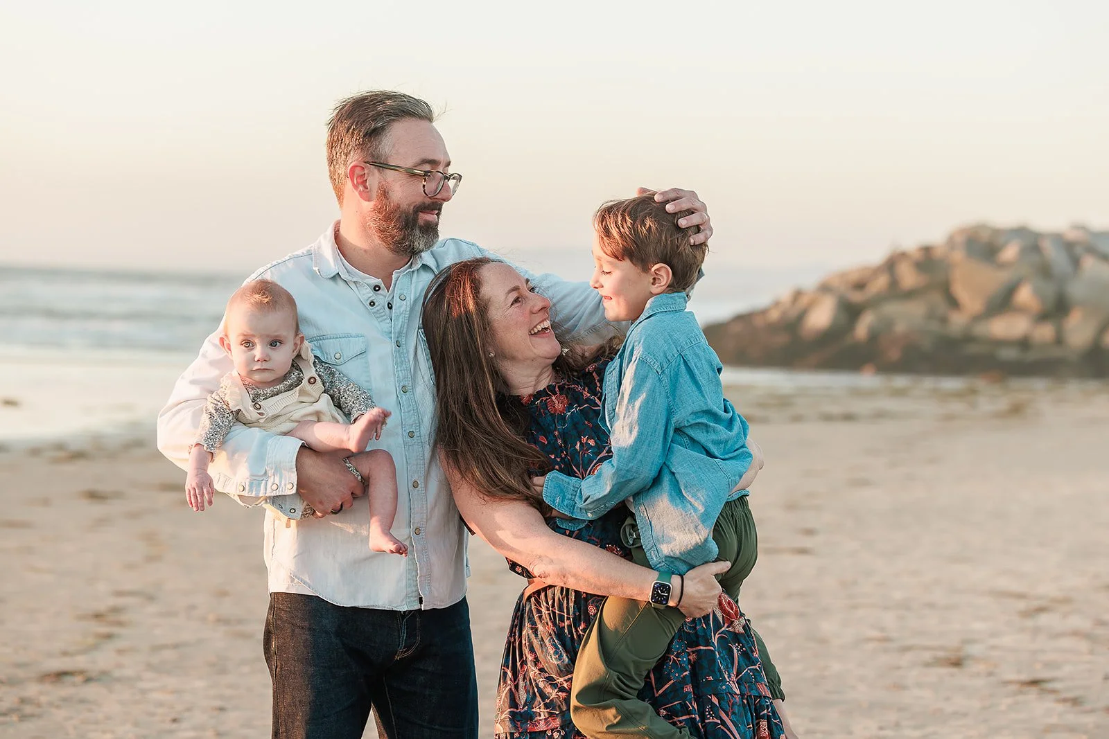 A family of four enjoying time together on the beach at sunset. The mother holds a young son, and the father holds a baby girl. They are smiling and looking at each other.