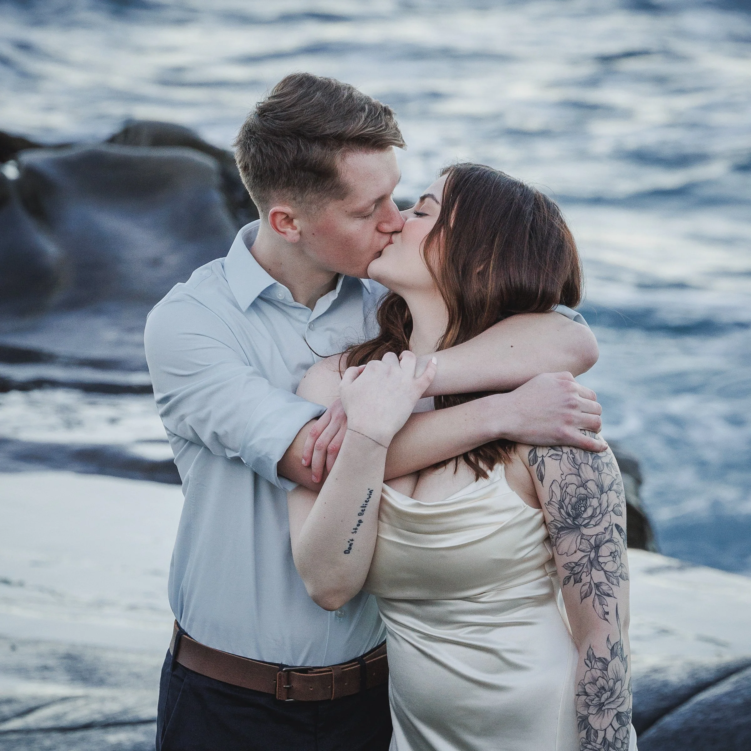 A young couple sharing a kiss by the ocean, with rocky shoreline in the background.