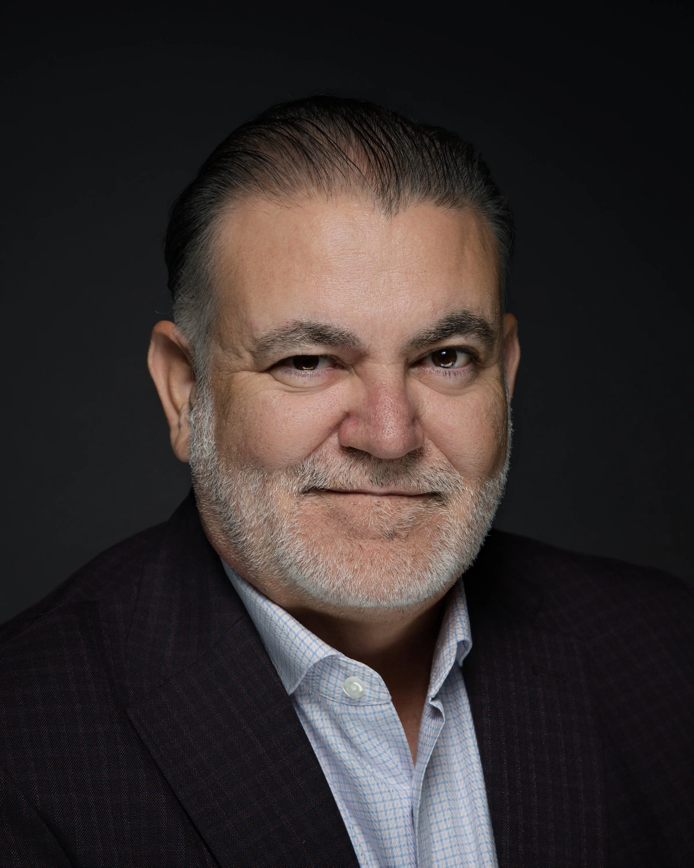 A middle-aged man with gray hair and beard, wearing a dark blazer and a light-colored shirt, smiling at the camera against a dark background.