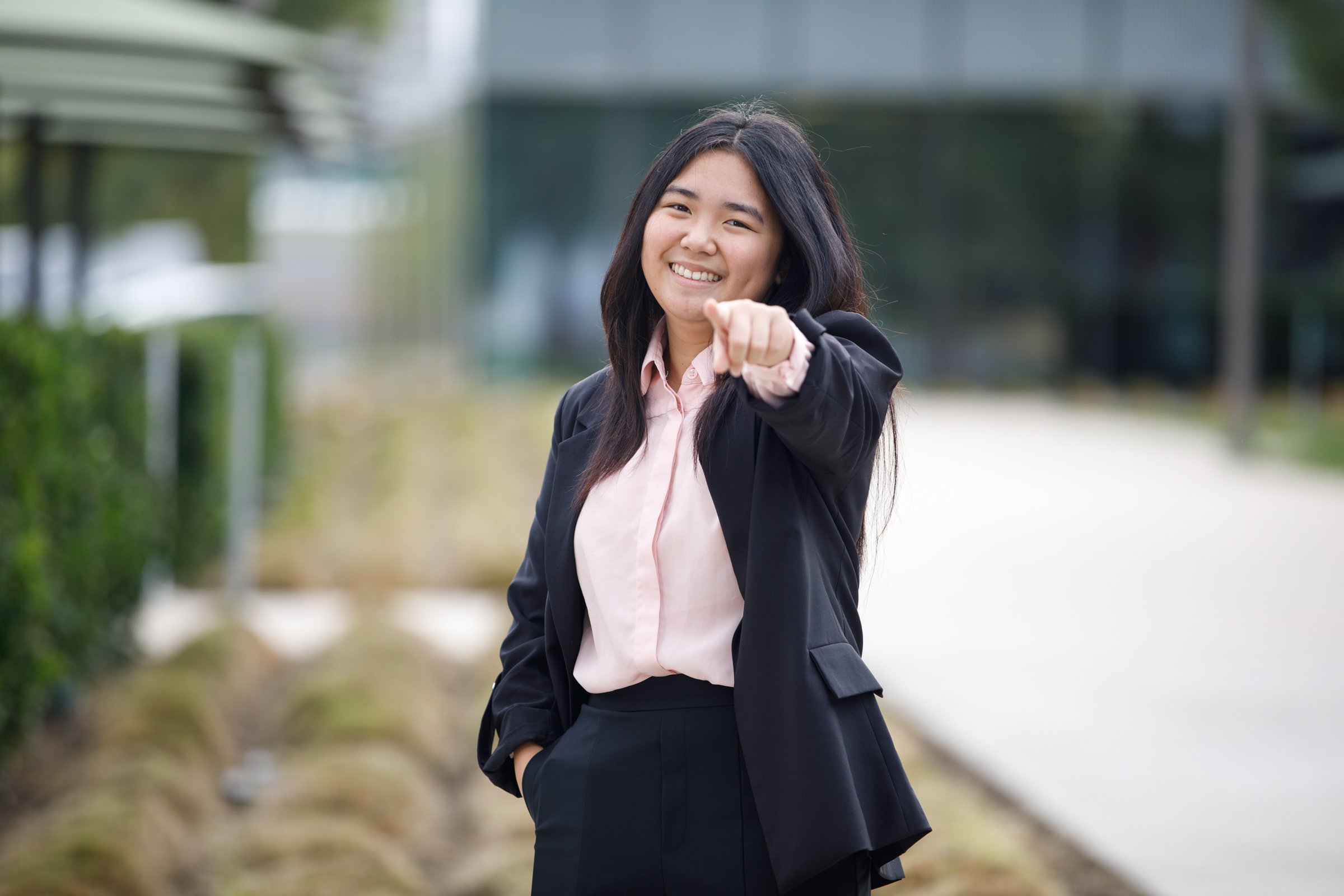 A young woman outdoors wearing a black blazer and light pink shirt, smiling and pointing at the camera.