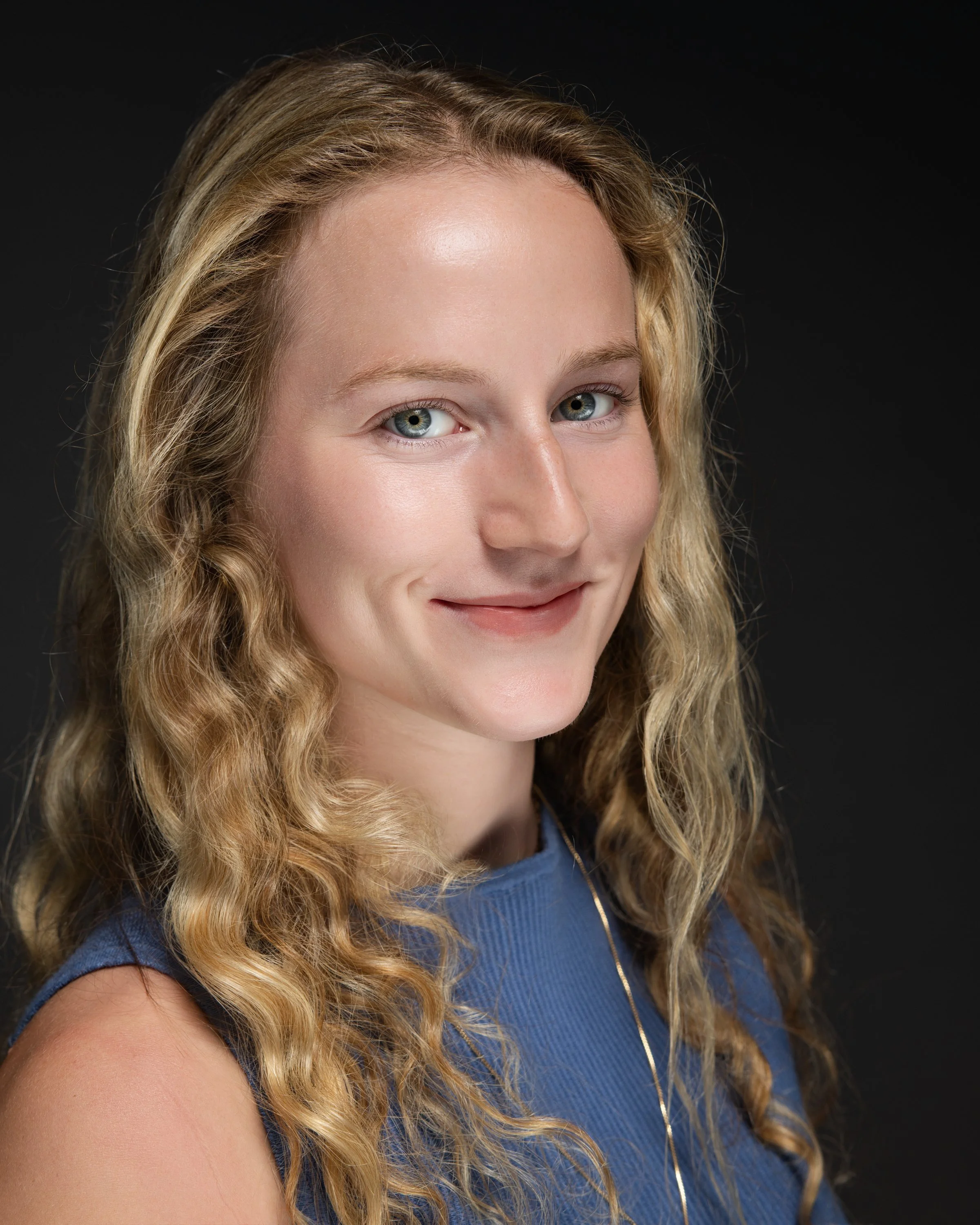 A portrait of a smiling woman with long, curly blonde hair, wearing a blue top and a gold necklace, against a dark background.