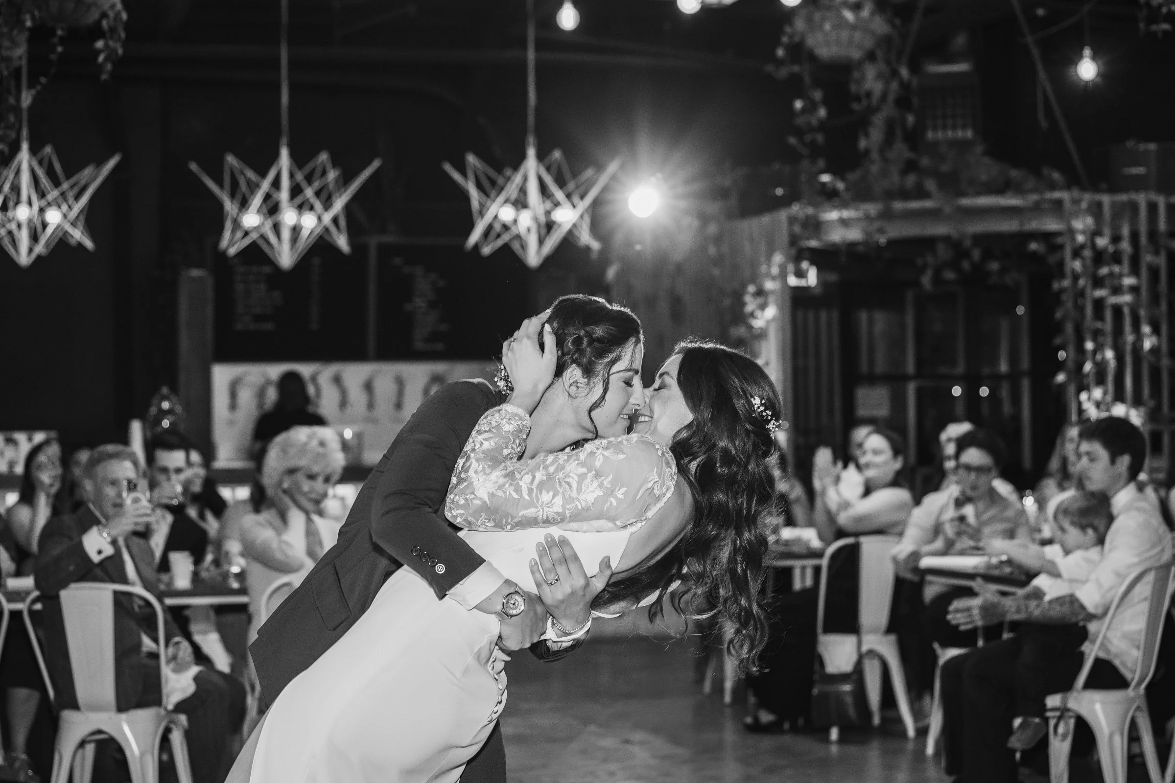 Black and white photo of a wedding reception showing a couple sharing a dance, surrounded by seated guests watching and taking photos.