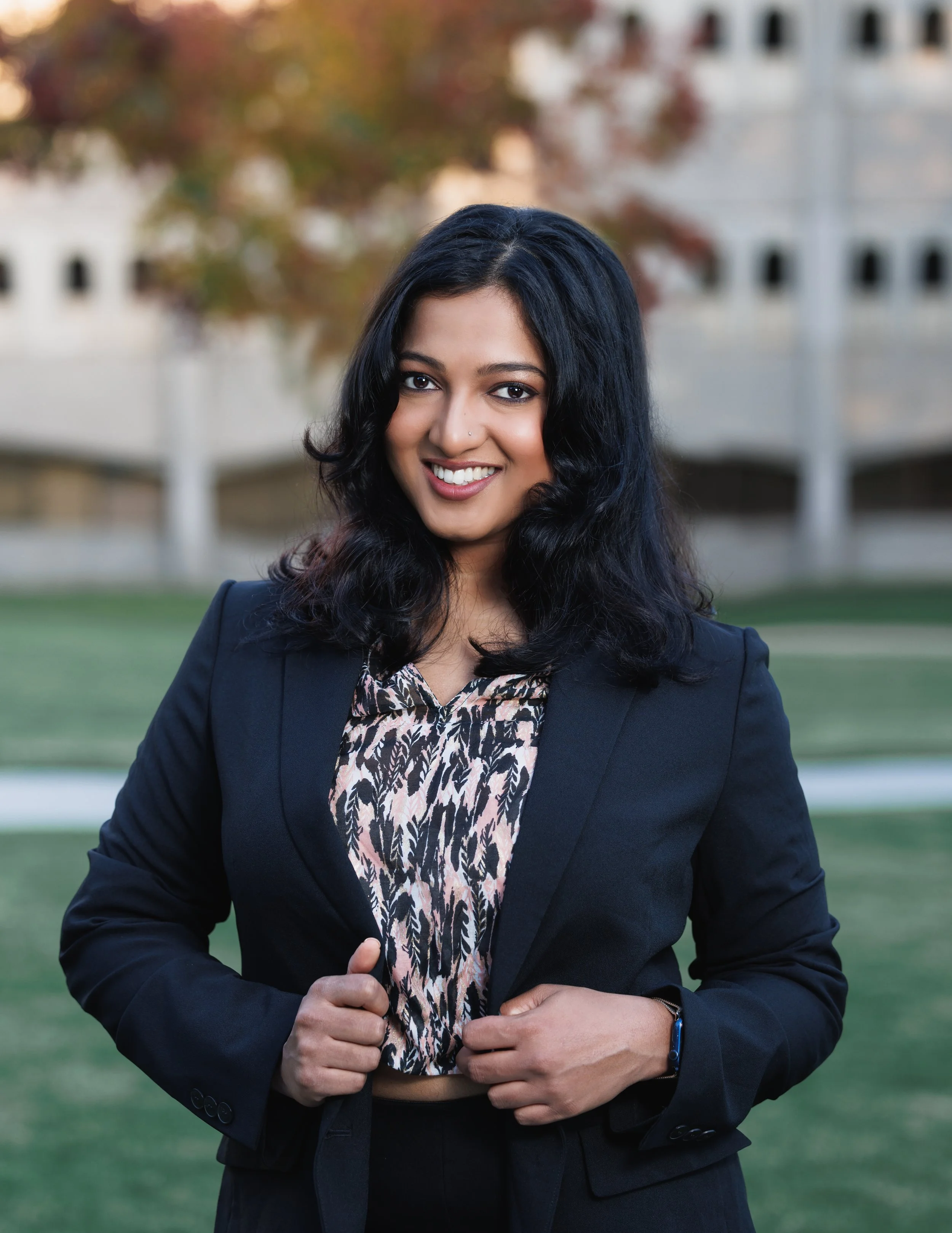 A woman with black hair smiling outdoors in a business suit, holding the edges of her jacket.