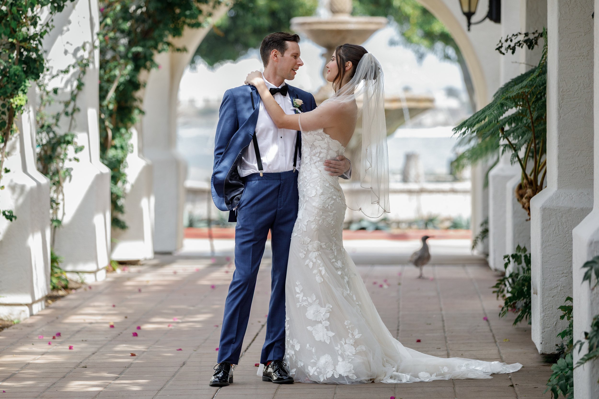 A bride and groom sharing a dance outdoors, with the groom holding his jacket and wearing a blue suit, and the bride in a white lace wedding gown and veil. They are smiling at each other under an arched walkway with greenery and a fountain in the bac