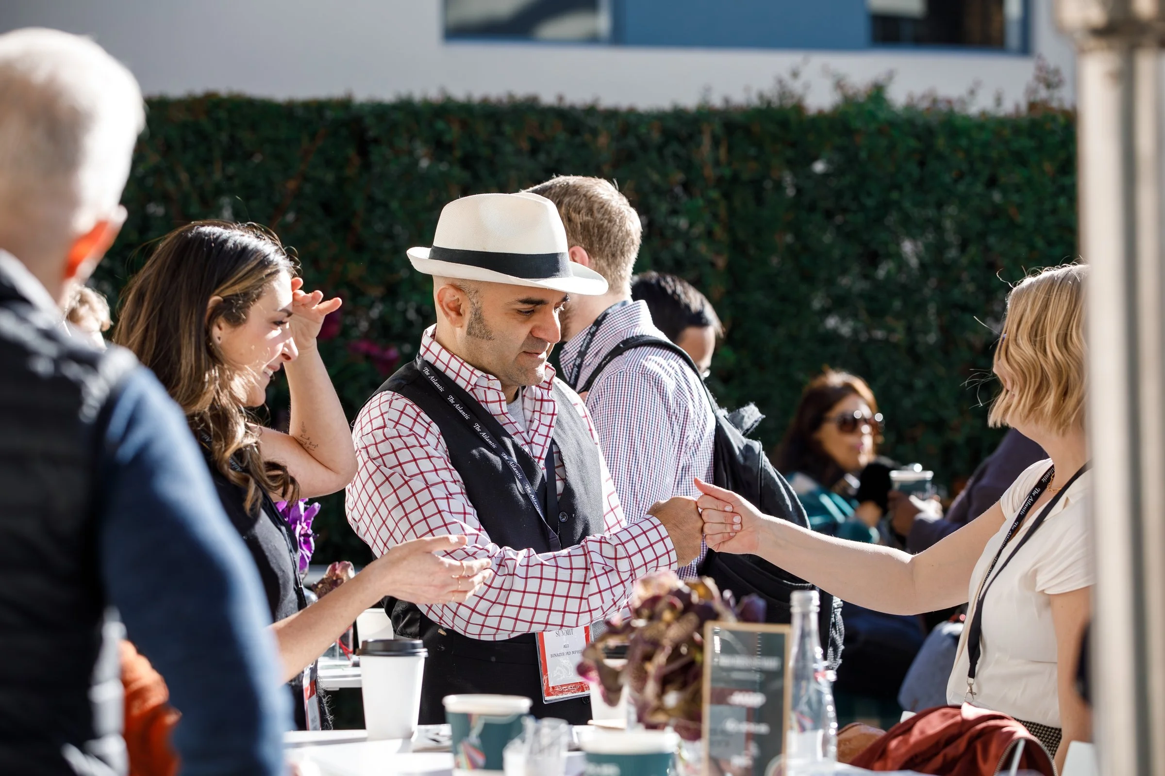 A man wearing a white fedora hat and checked shirt shaking hands with a woman at an outdoor event, with several people standing nearby.