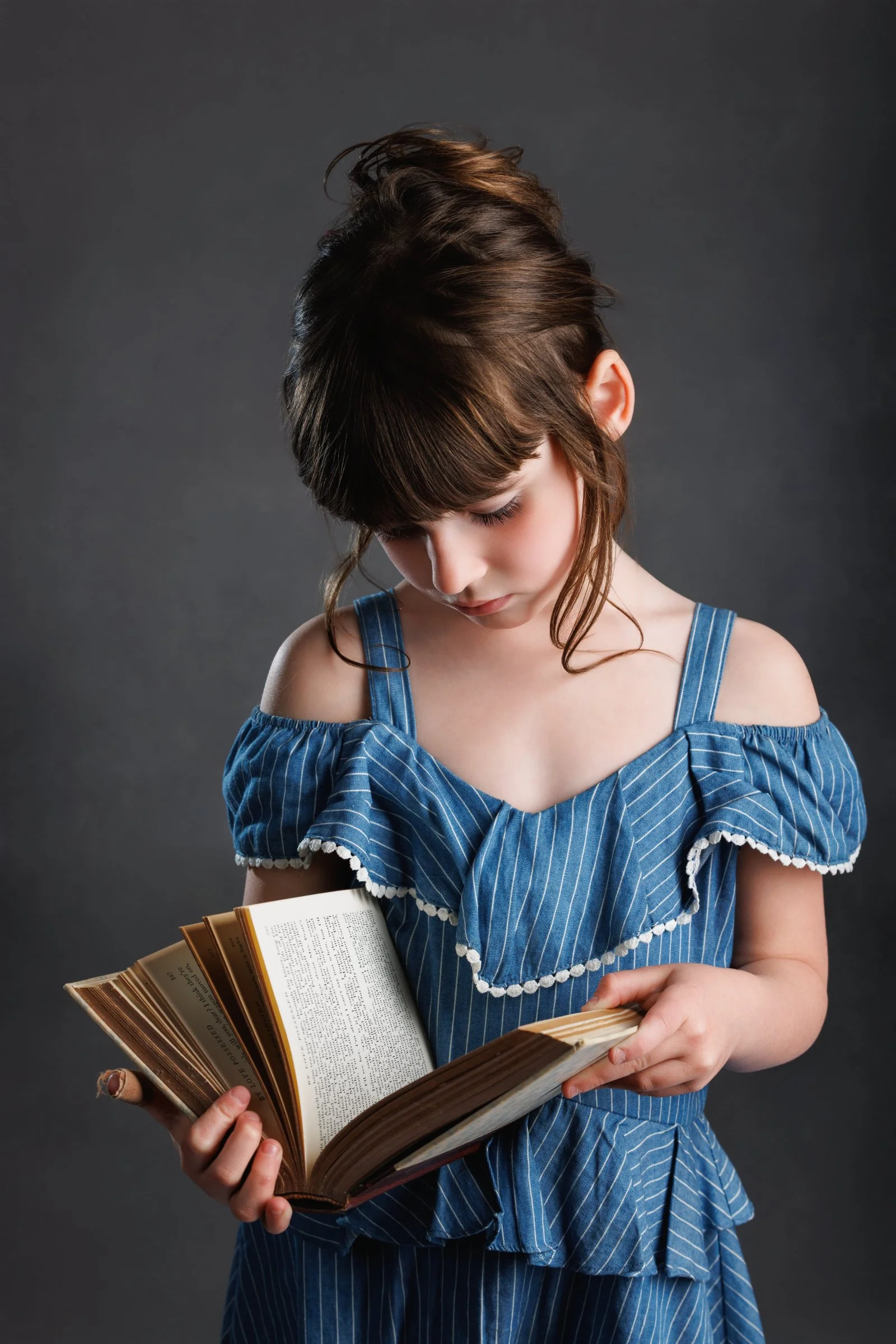 A young girl with brown hair and bangs, wearing a blue off-shoulder dress with white trim, is looking down at an open book she is holding in both hands, against a dark gray background.