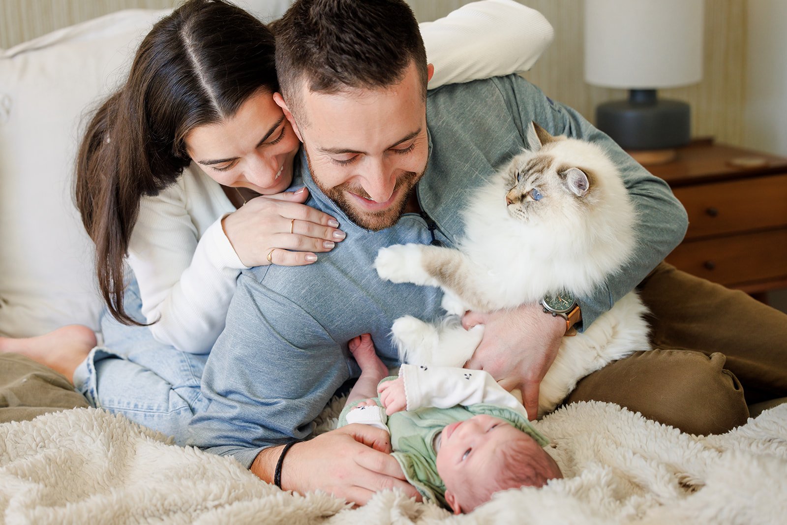 A happy couple plays with their baby and cat on a bed in a cozy bedroom.