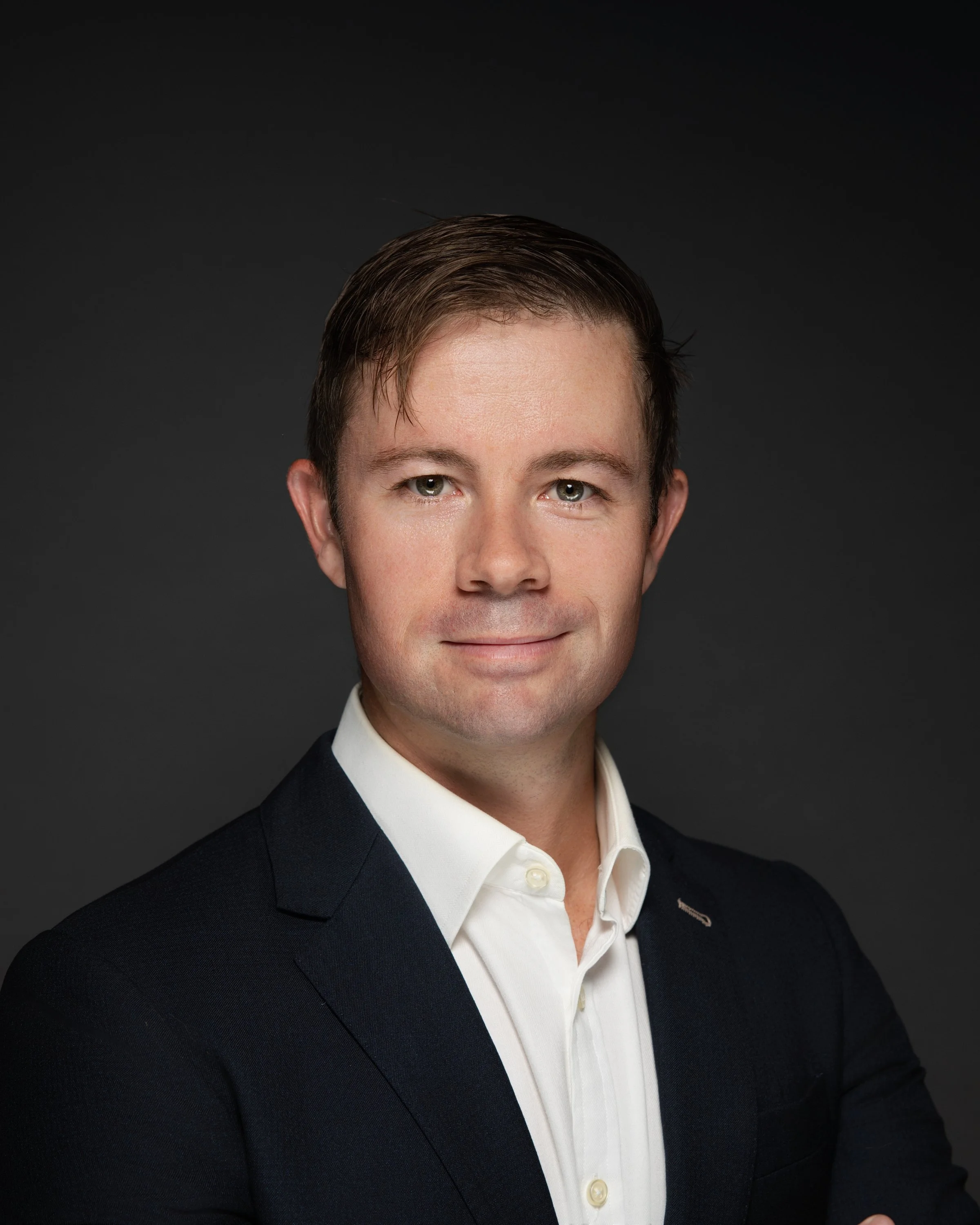 Professional headshot of a man in a dark suit and white shirt, smiling slightly, against a dark background.