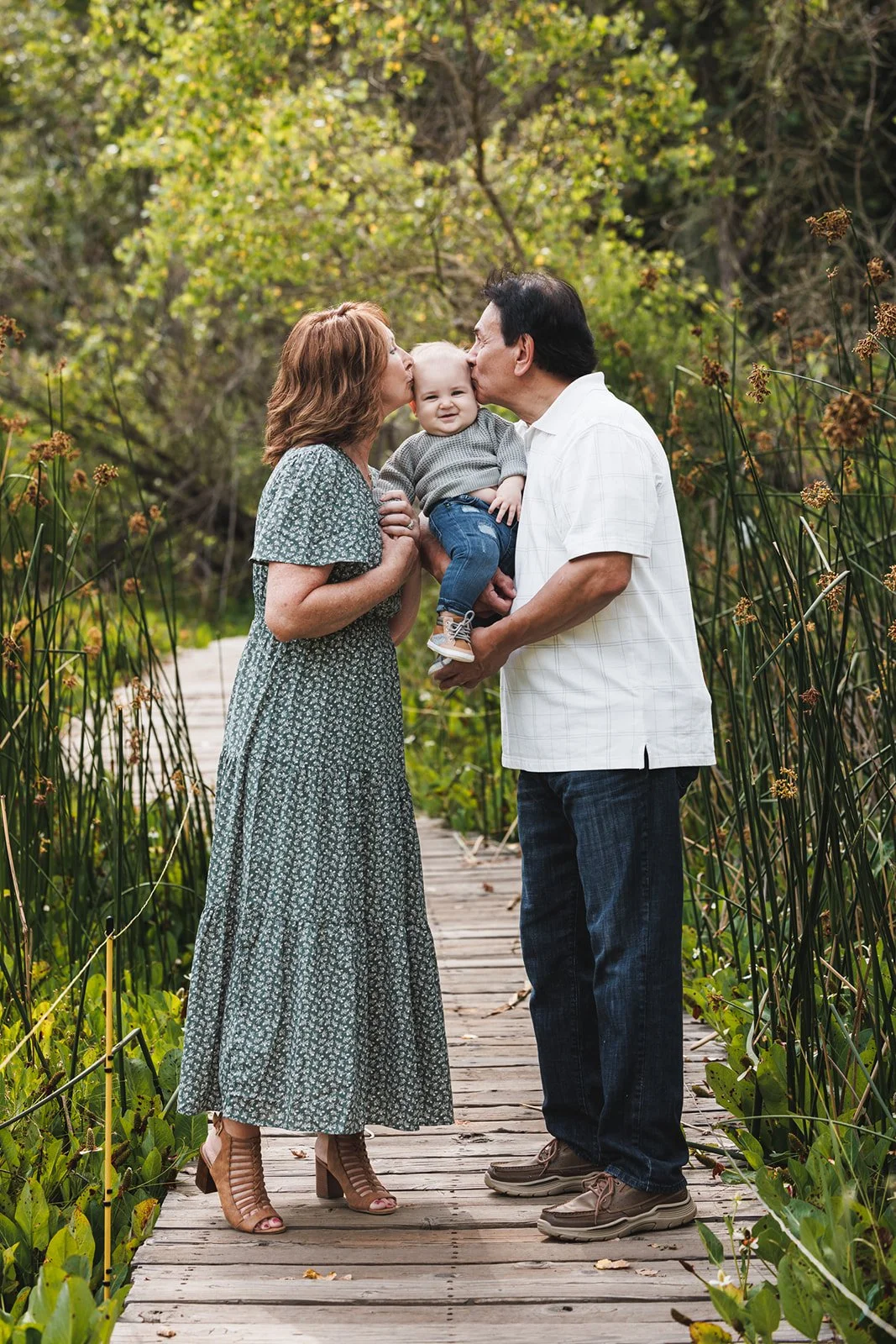 A family of three standing on a wooden dock surrounded by greenery, with the mother and father kissing their smiling young child on the cheeks.
