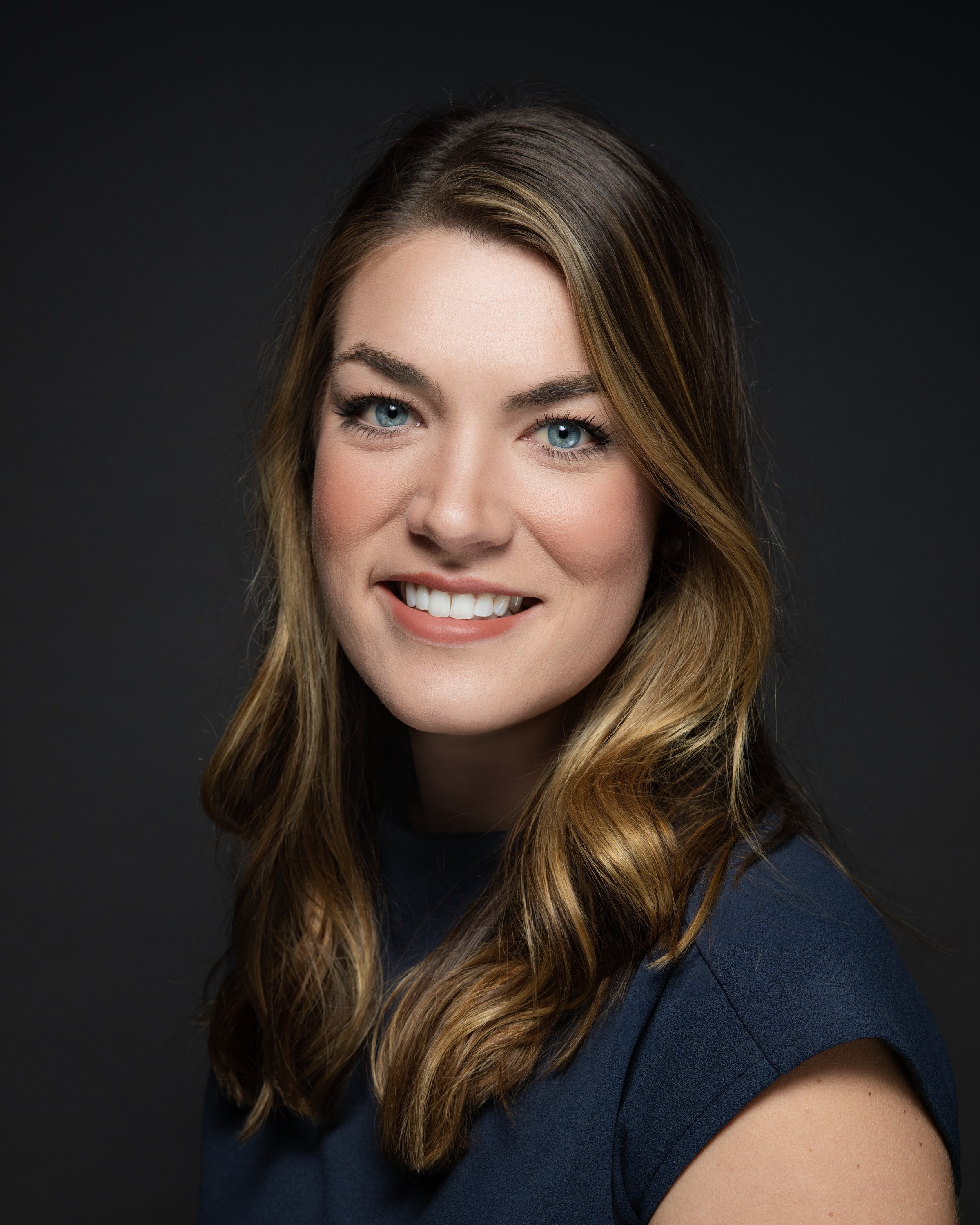 Portrait of a young woman with long, wavy brown hair, blue eyes, and fair skin, smiling against a dark background.