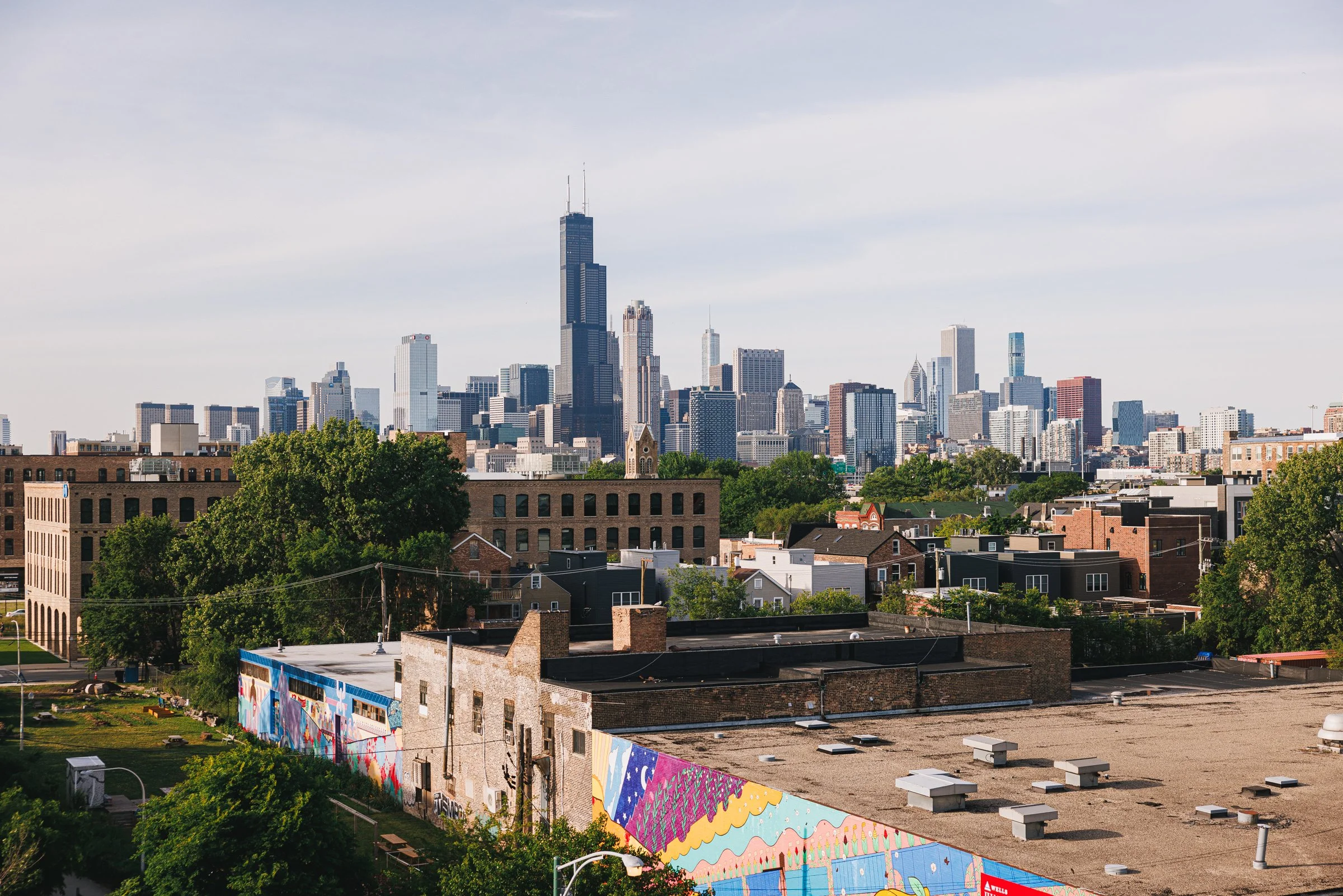 City skyline with tall skyscrapers, including the Willis Tower, viewed from a residential area with trees, buildings, and a colorful mural on a wall in the foreground.