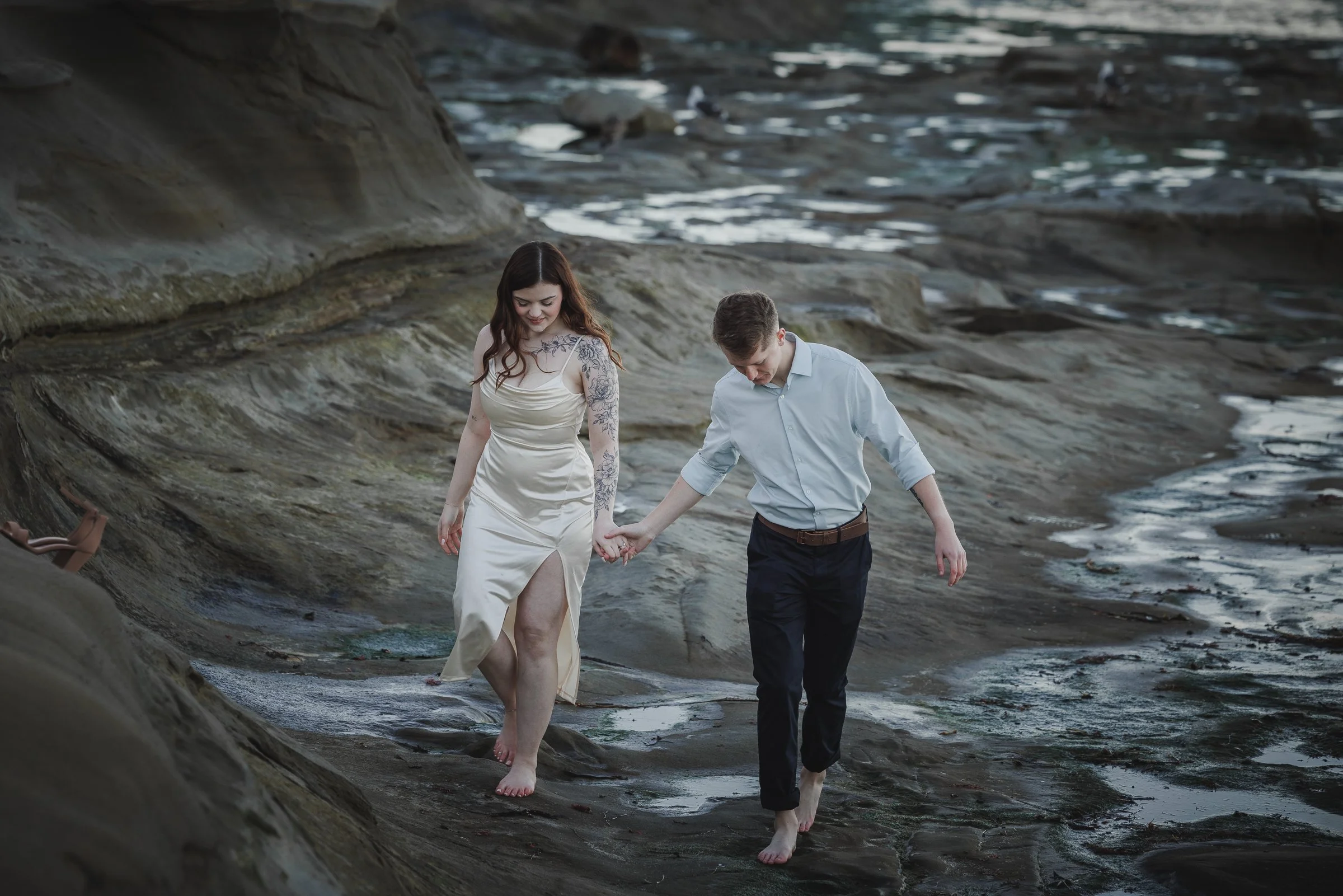 A couple walking barefoot along a rocky shoreline holding hands.