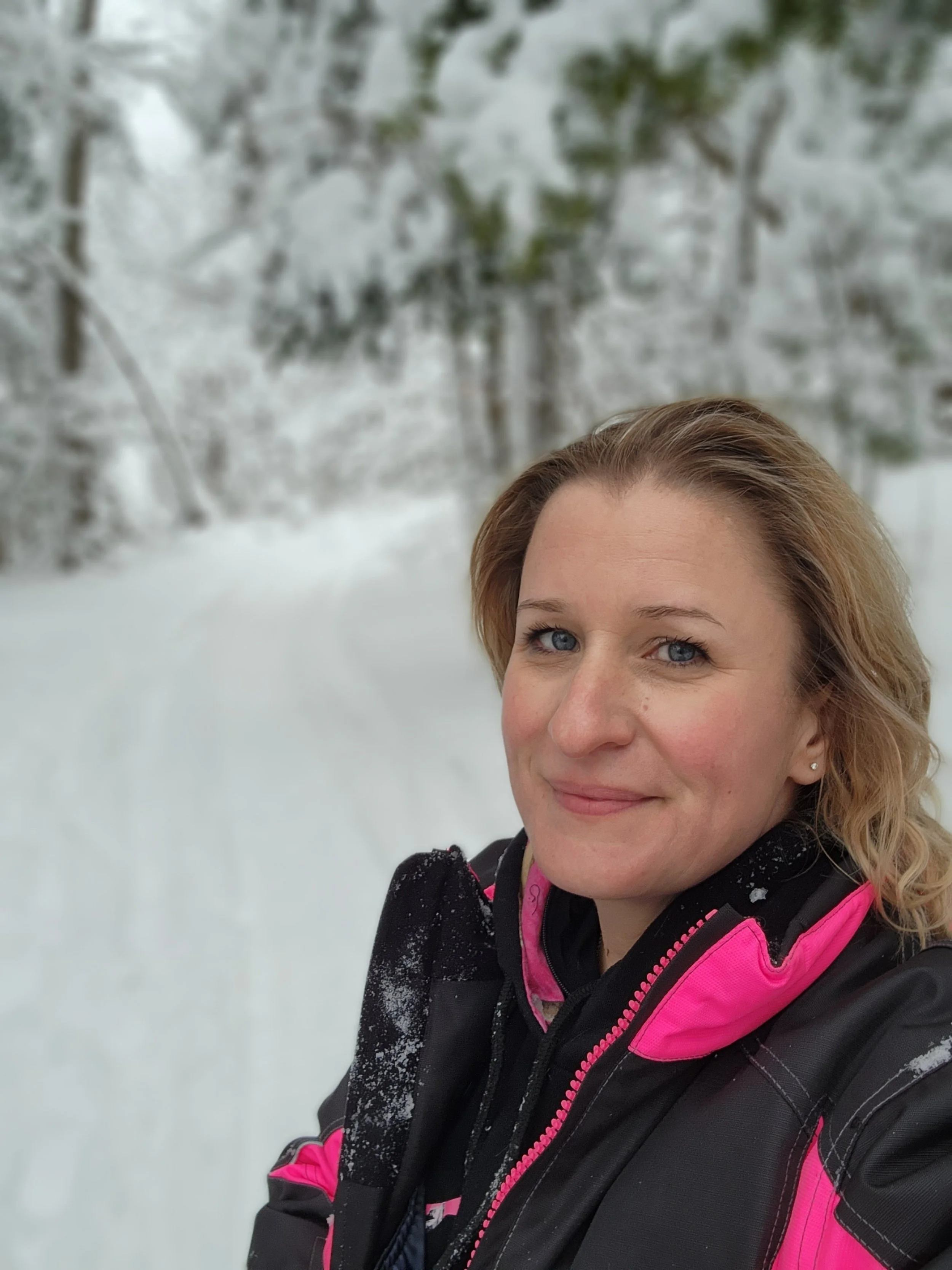 The owner of Retained Legal Services. She has blonde, wavy hair and blue eyes taking a selfie in a snowy, forested area, wearing a black and pink snowmobile jacket.