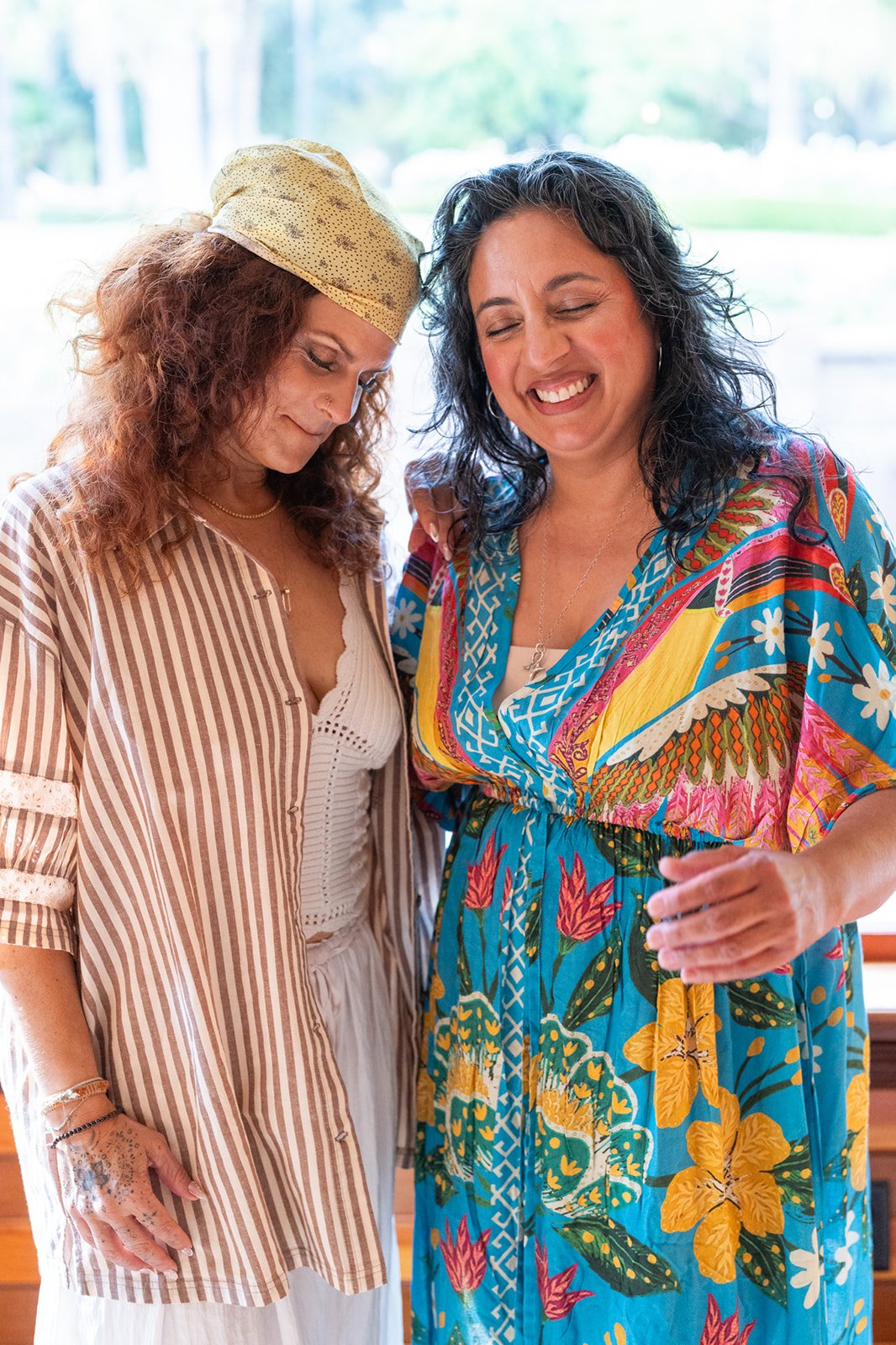 Two women, one with curly hair wearing a colorful boho dress, and the other with long curly hair wearing a striped shirt and headscarf, are smiling and sharing a moment together indoors.