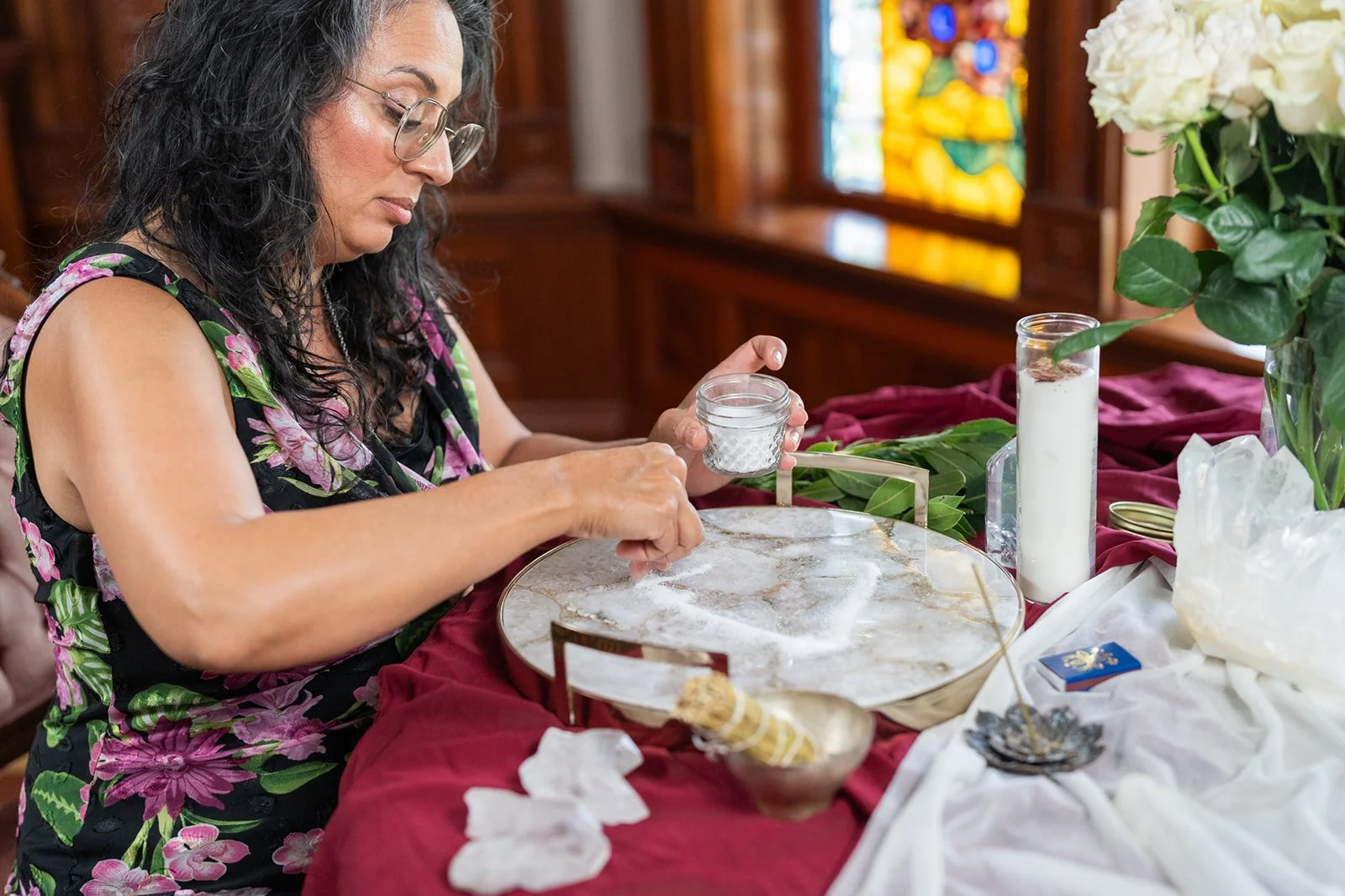 Woman in glasses placing a small container of salt or sugar on a decorated table with flowers, candles, and crystals.