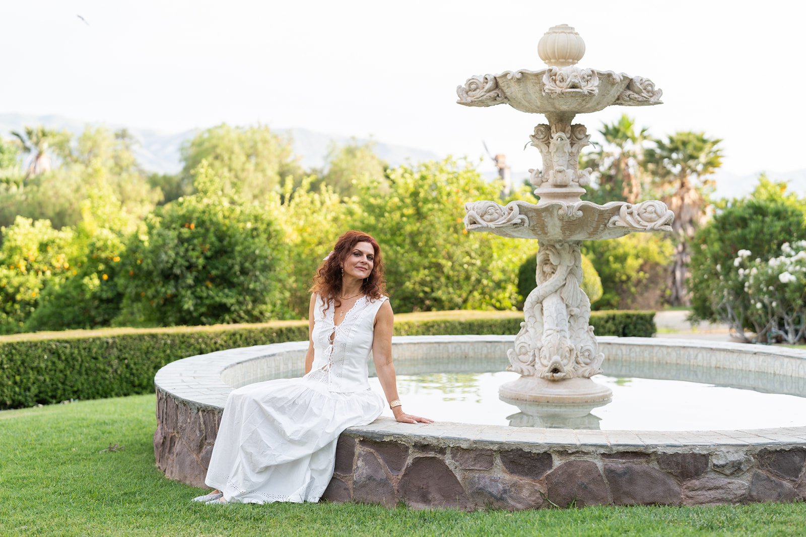 A woman with curly red hair in a white dress sitting by a stone fountain with dragon sculptures in a garden with green trees and bushes.