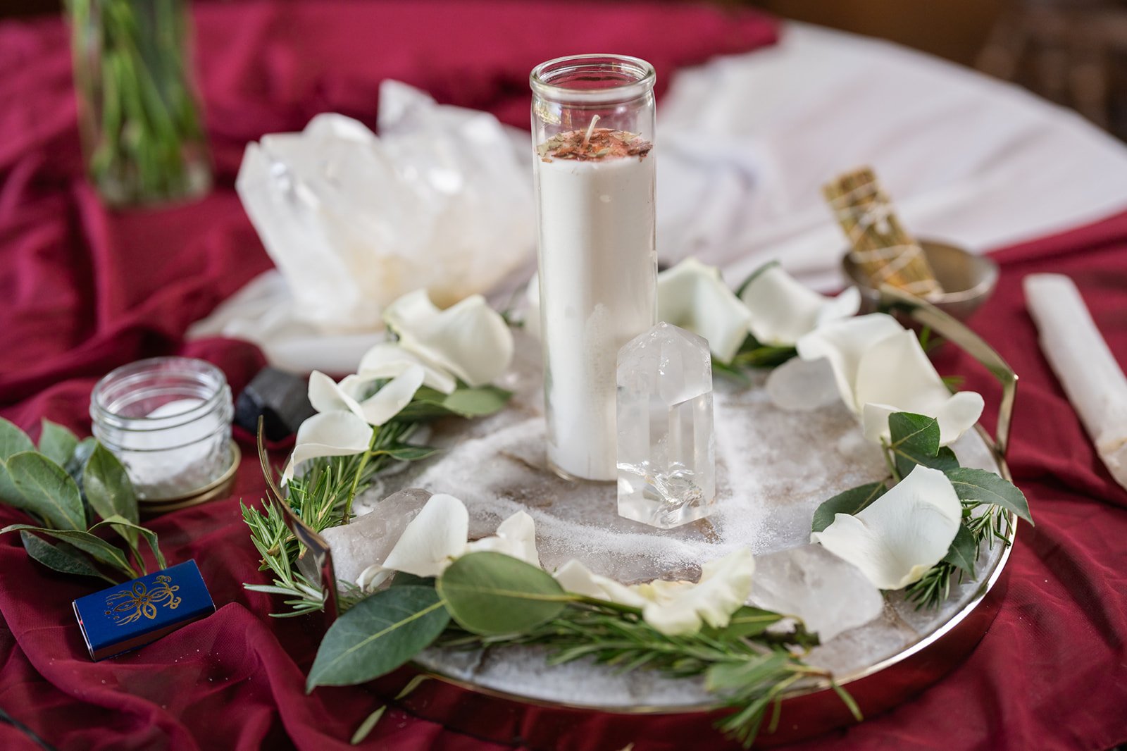 A memorial or remembrance display with a candle, large clear quartz crystal, and white flowers on a marble tray, surrounded by red fabric.