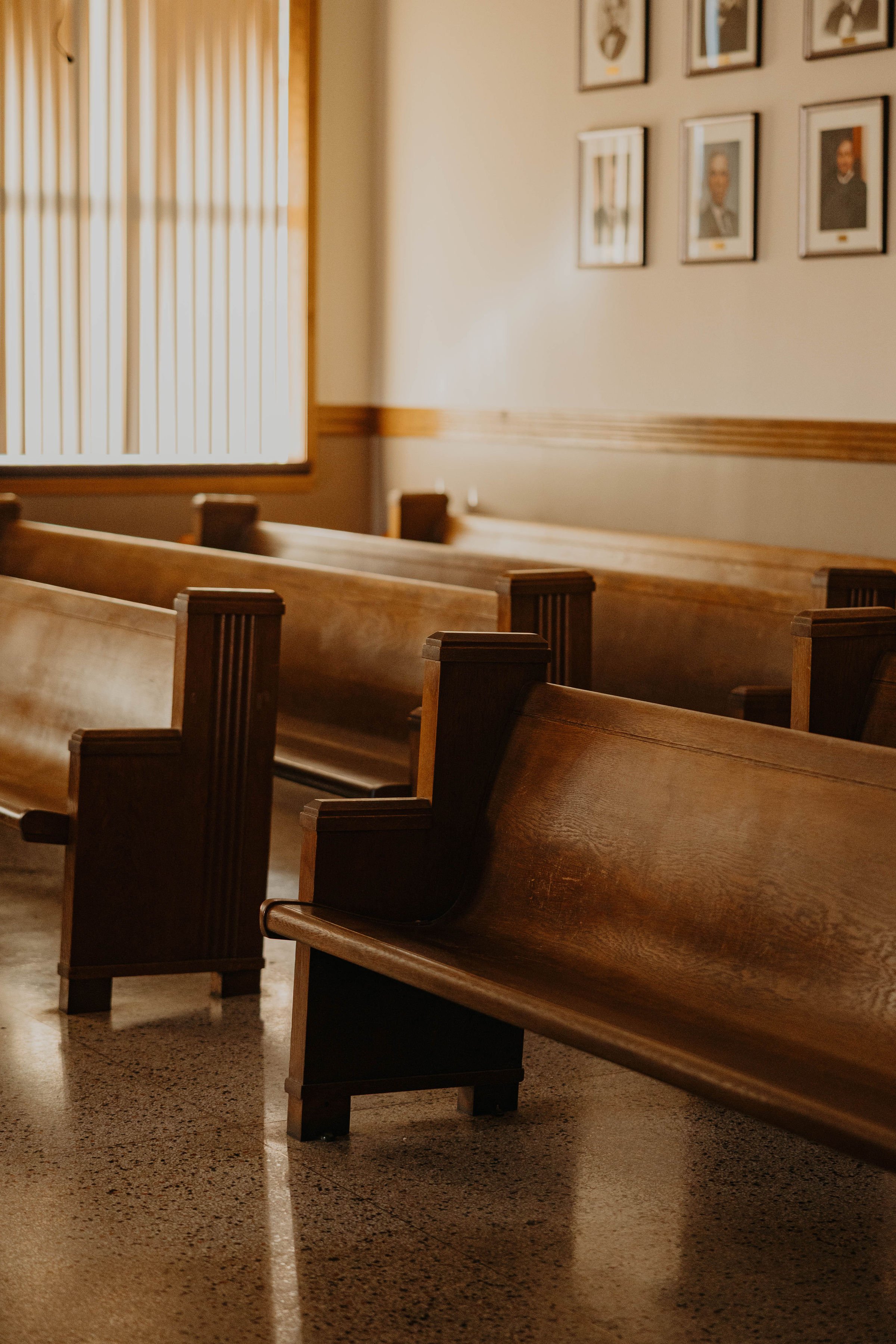 Empty church pews inside a chapel with framed photographs on the wall and a window with curtains letting in natural light.
