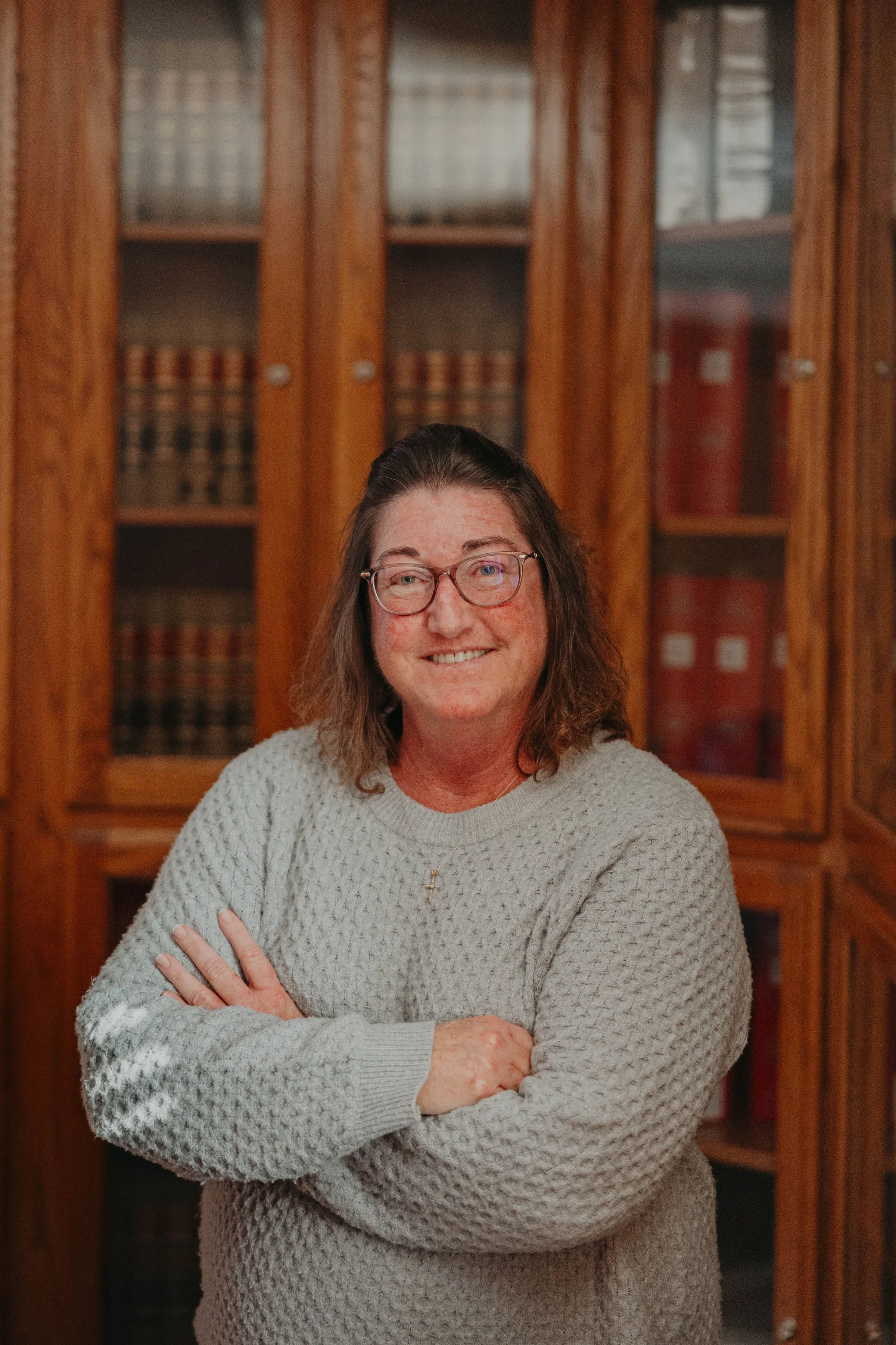 A woman with long brown hair, glasses, and a cross necklace wearing a gray knitted sweater, standing with her arms crossed in front of a wooden bookshelf with glass doors filled with books.
